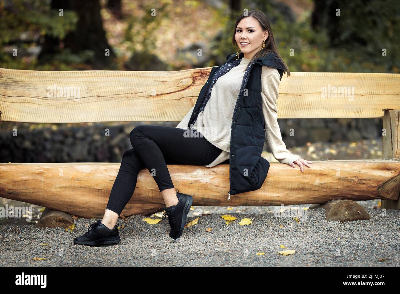 Ritratto autunnale di giovane donna asiatica con capelli marroni in abiti di colore base in parco su panca di legno all'aperto Foto Stock