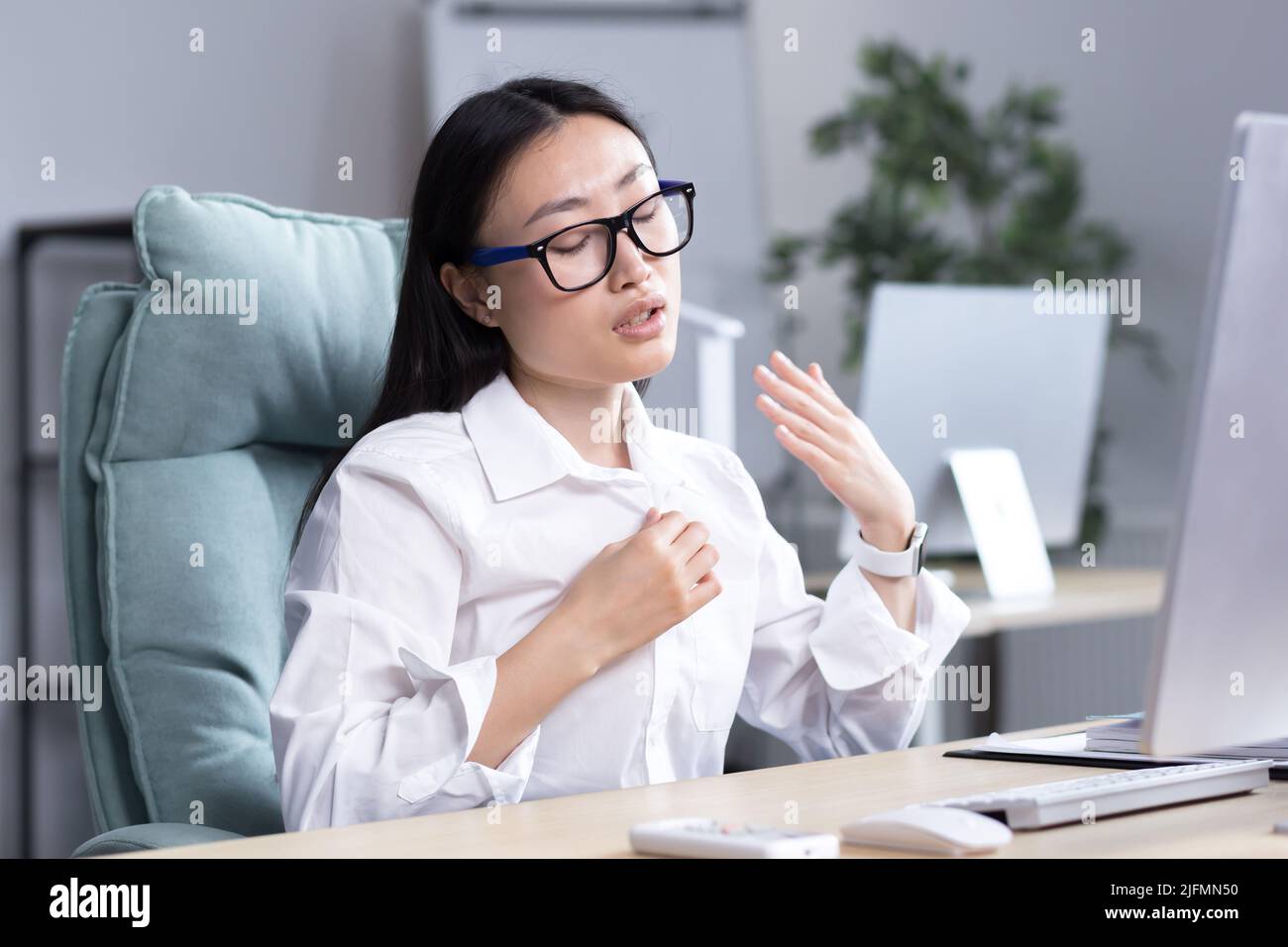 Calore in ufficio, donna asiatica che cerca di rinfrescarsi, donna d'affari che lavora al computer, agitando le mani per raffreddarsi. Foto Stock