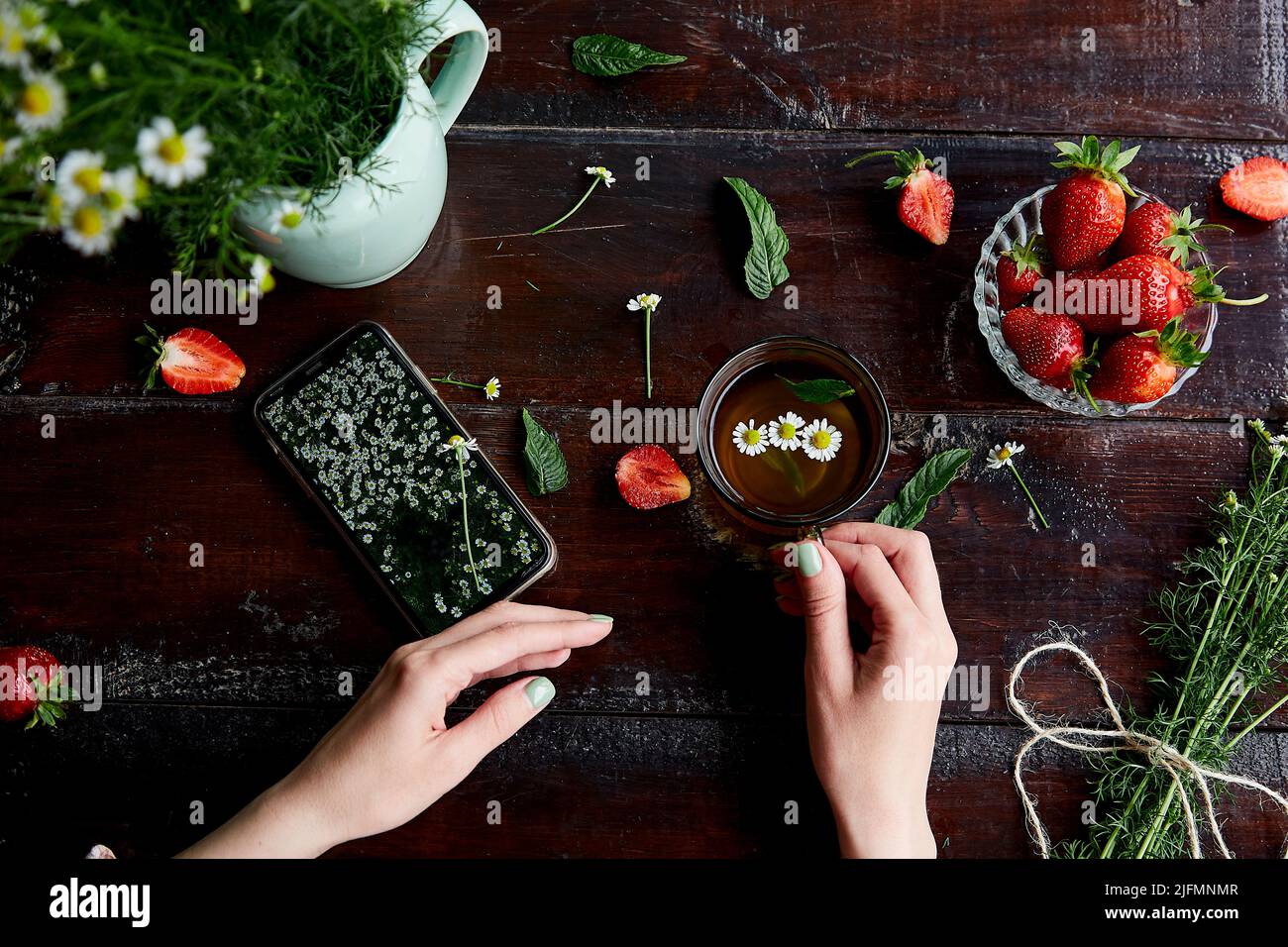 Donna mani che tengono tazza di tè camomilla e telefono cellulare. D'estate, fragole fresche, camomilla, tè naturale e menta. Mock up di schermo mobile con campo di fiori camomilla. Foto Stock