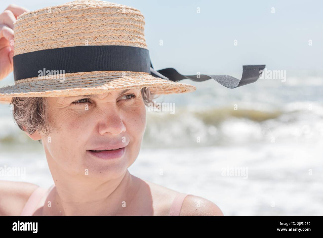 Una donna leggermente sorridente dai capelli grigi che tiene il boater dal vento di mare che fluttering nastro nero sul suo cappello contro sfondo sfocato di mare con grandi onde. Foto Stock