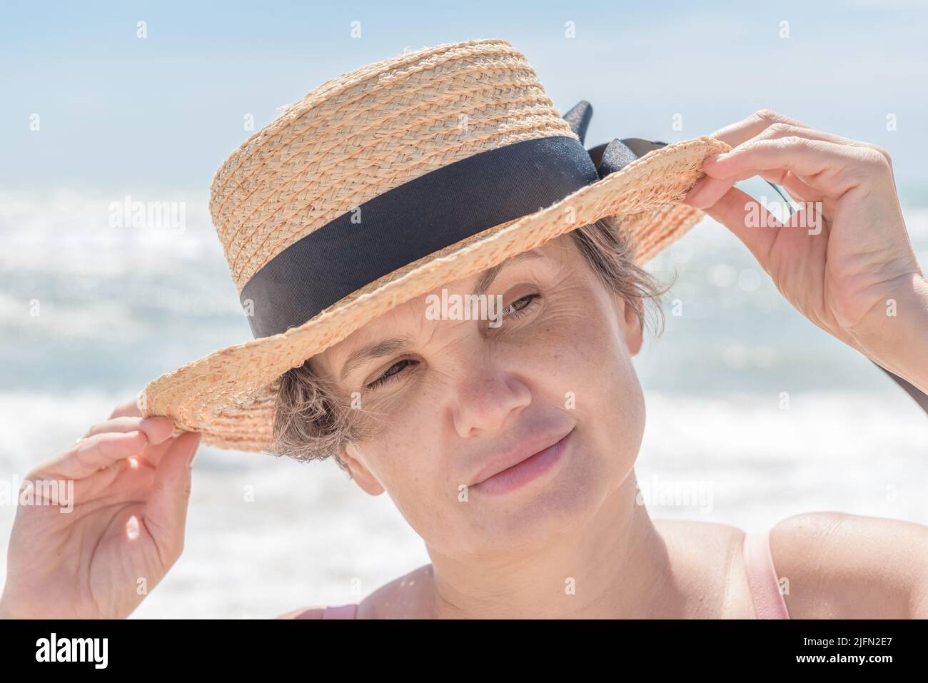 Ritratto di donna dai capelli grigi che sostiene le brime del suo boater dalla brezza marina con entrambe le mani, guardando via con un piccolo sorriso.concetto di femminilità. Foto Stock