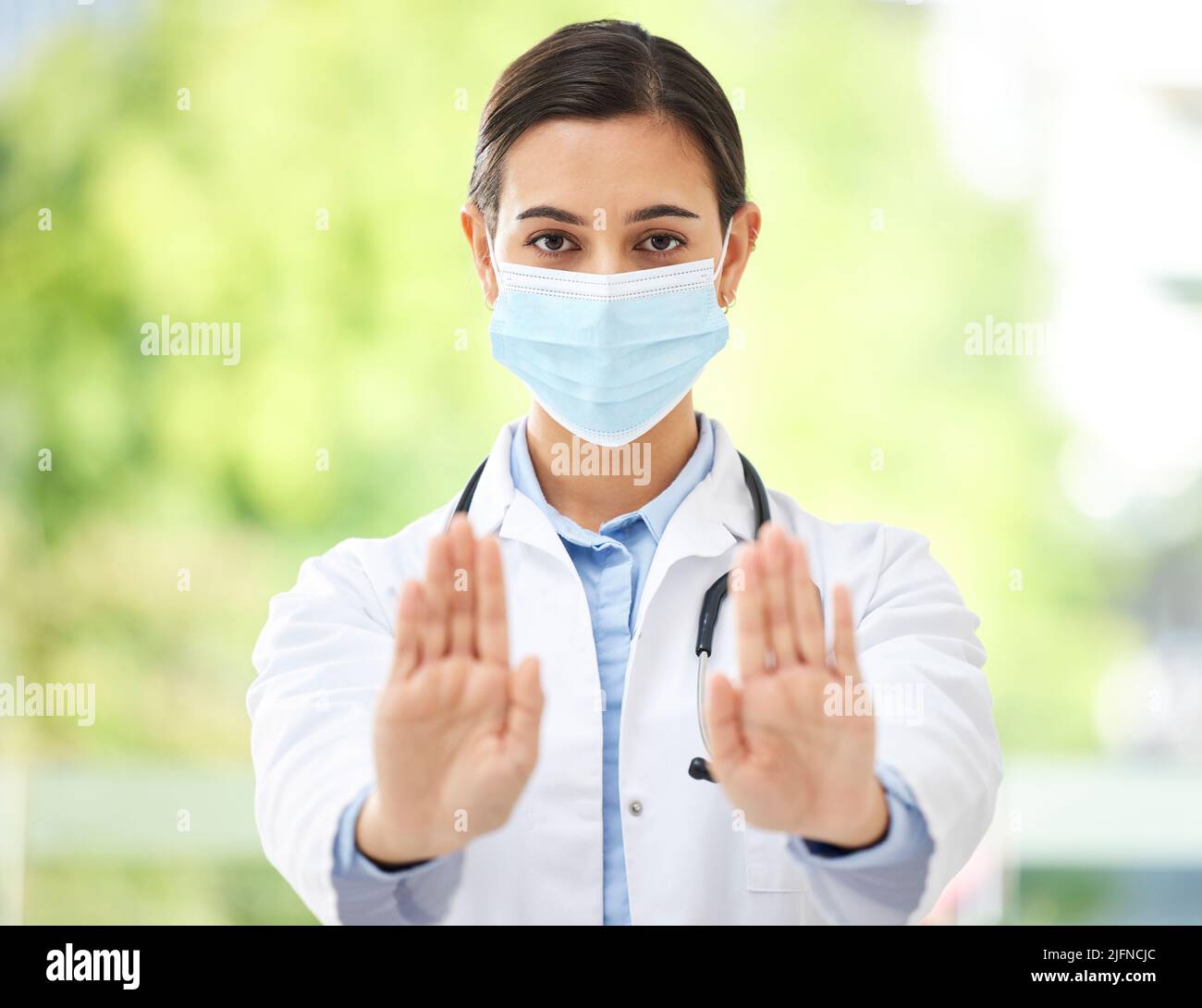 Ritratto di una donna ispanica medico che indossa una maschera e mostra un gesto di arresto con la mano mentre indossa uno stetoscopio in un ufficio ospedaliero. Foto Stock