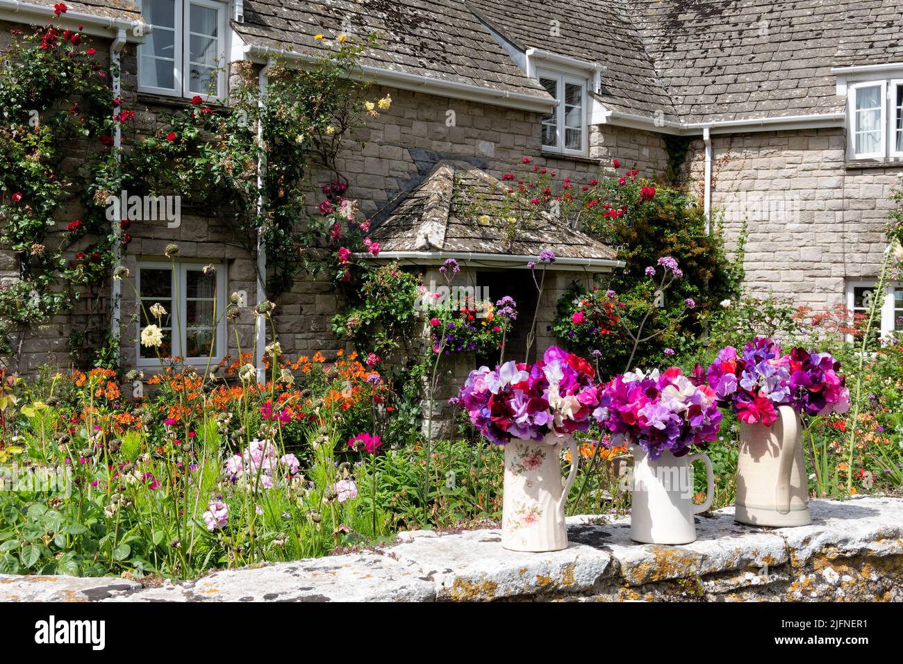 Un grazioso giardino cottage inglese in pieno fiore in una giornata di sole. Fiori recisi in caraffe sono posti sulla parete del giardino a sfondo di piante tradizionali Foto Stock