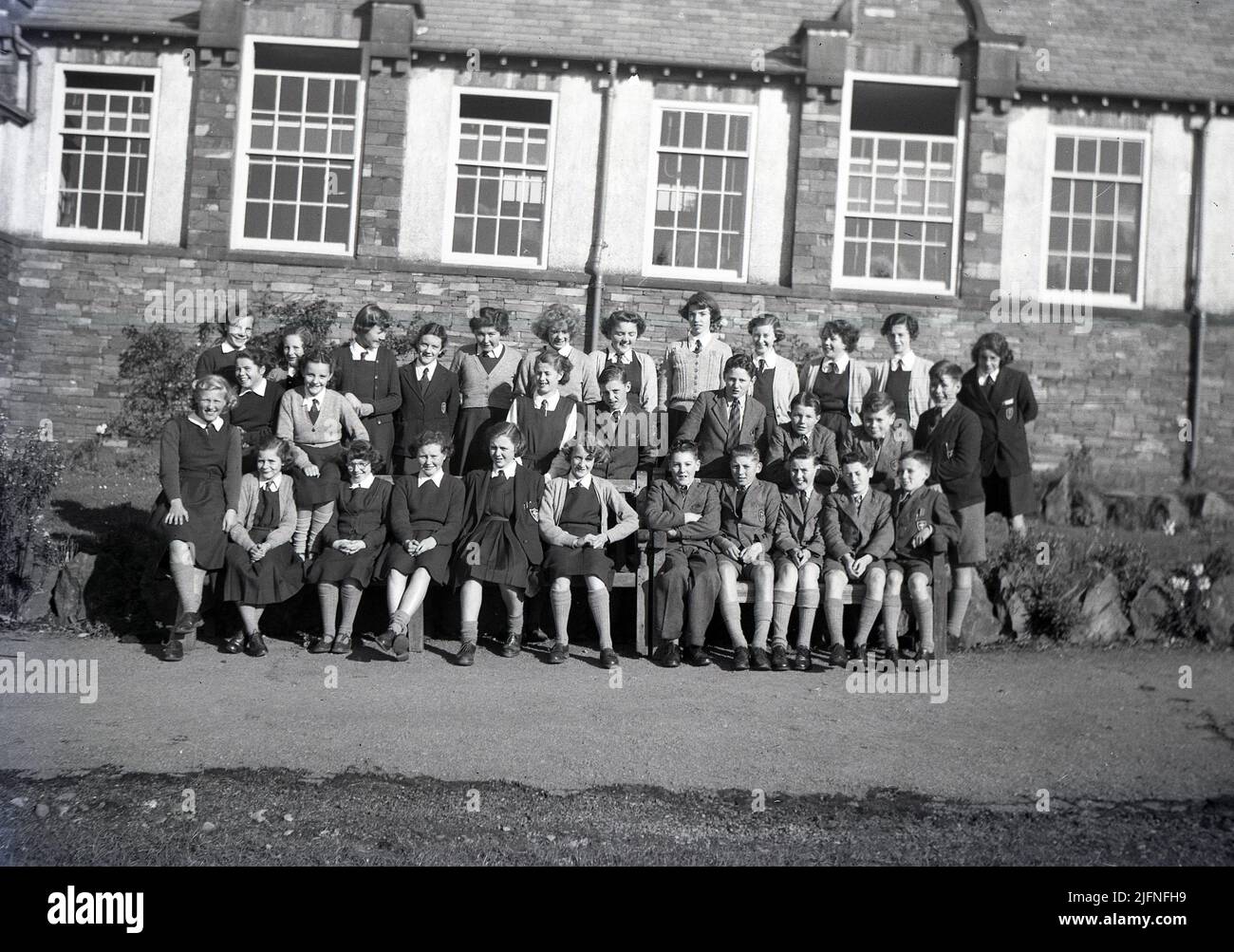 1951, storico, una grande classe di bambini eccitati nella loro uniforme, seduti e in piedi insieme fuori dal loro edificio scolastico per una foto di classe di gruppo, Inghilterra, Regno Unito. Foto Stock