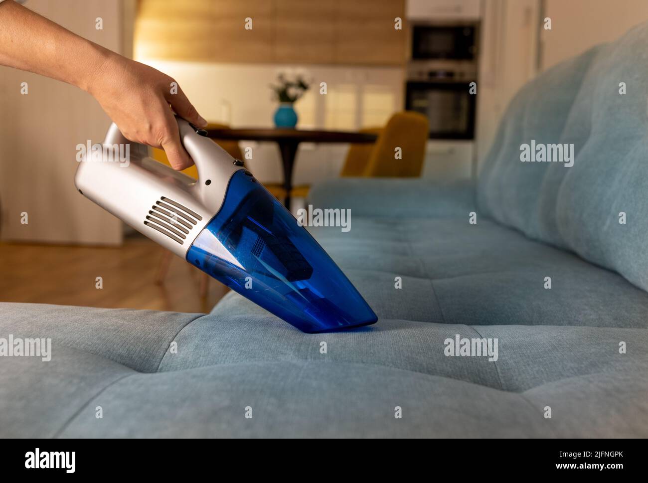 Primo piano della mano femminile con guanti che tengono comodo aspirapolvere wireless ricaricabile che si agganciano i capelli nad briciole dal divano in soggiorno con cucina A. Foto Stock