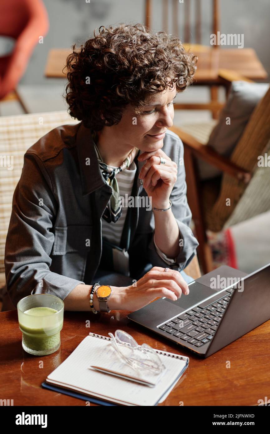 Felice uomo d'affari di mezza età o di rete freelance da tavolo in caffetteria mentre tiene la mano sul mento e guardando i dati sullo schermo Foto Stock