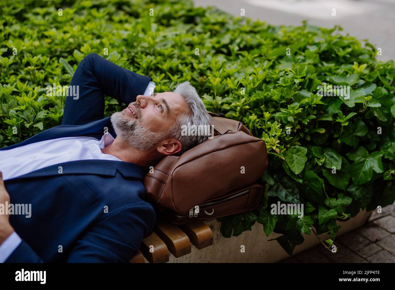 Uomo d'affari maturo che si rilassa su panchina nel parco cittadino durante la pausa al lavoro, concetto di equilibrio vita-lavoro. Foto Stock