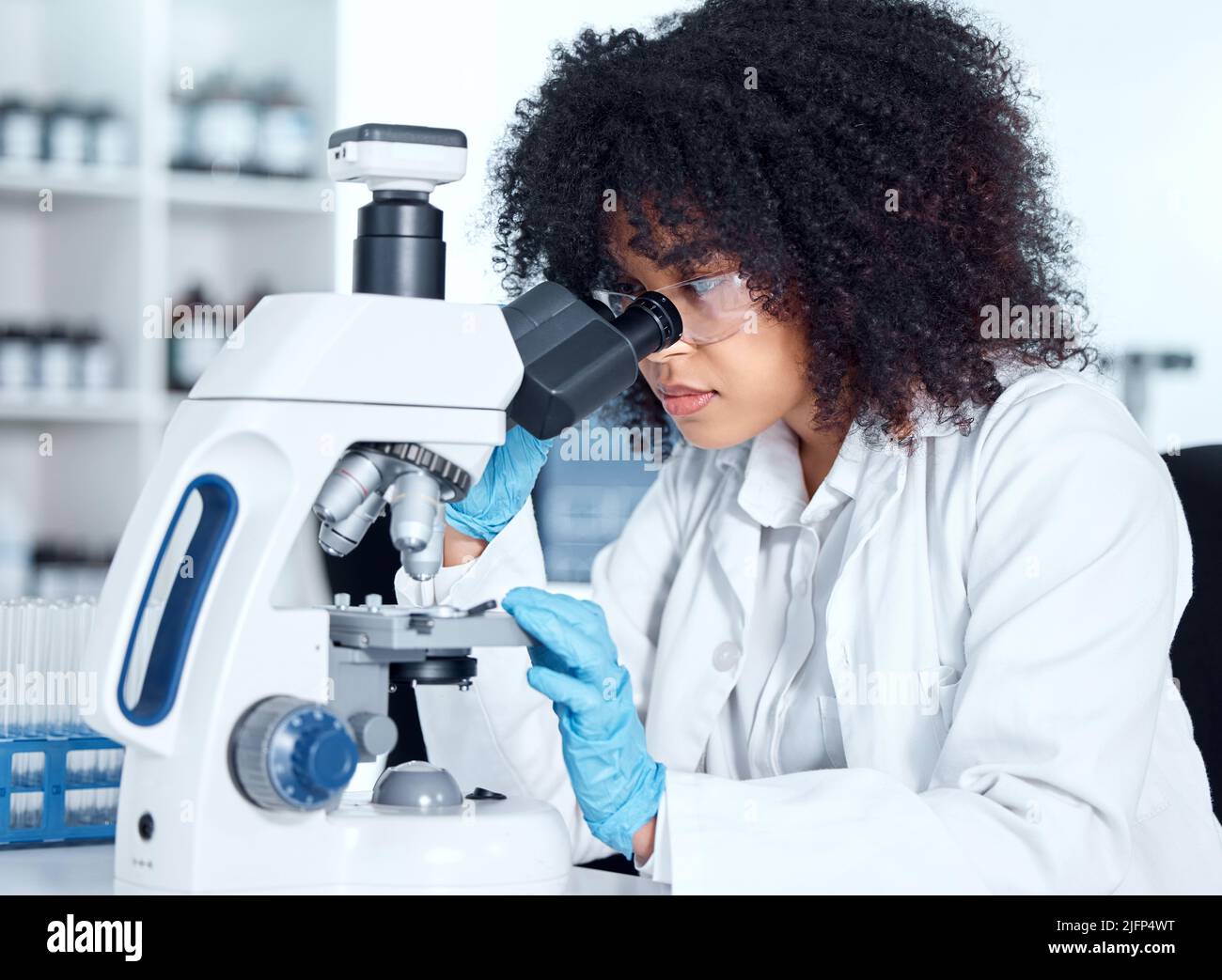 Giovane donna afroamericana che indossa un camice da laboratorio e occhiali che guarda campioni medici su un microscopio nel suo laboratorio. Una scienziata femminile mista Foto Stock