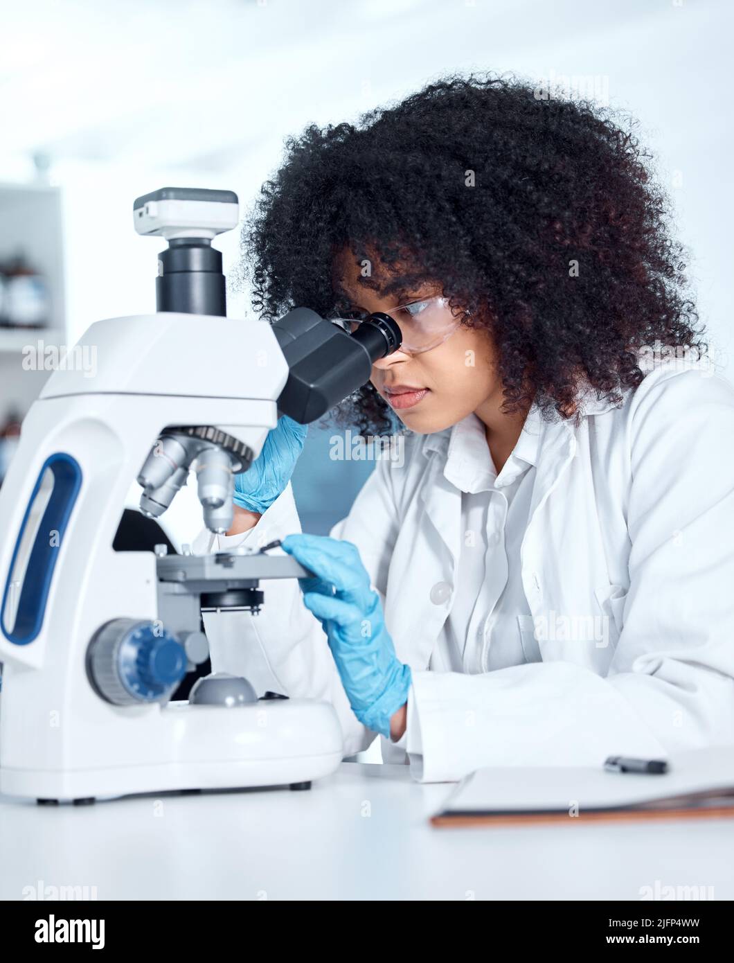 Una bella giovane donna afroamericana con un afro che indossa un camice da laboratorio e guarda campioni medici su un microscopio nel suo laboratorio. Una gara mista Foto Stock