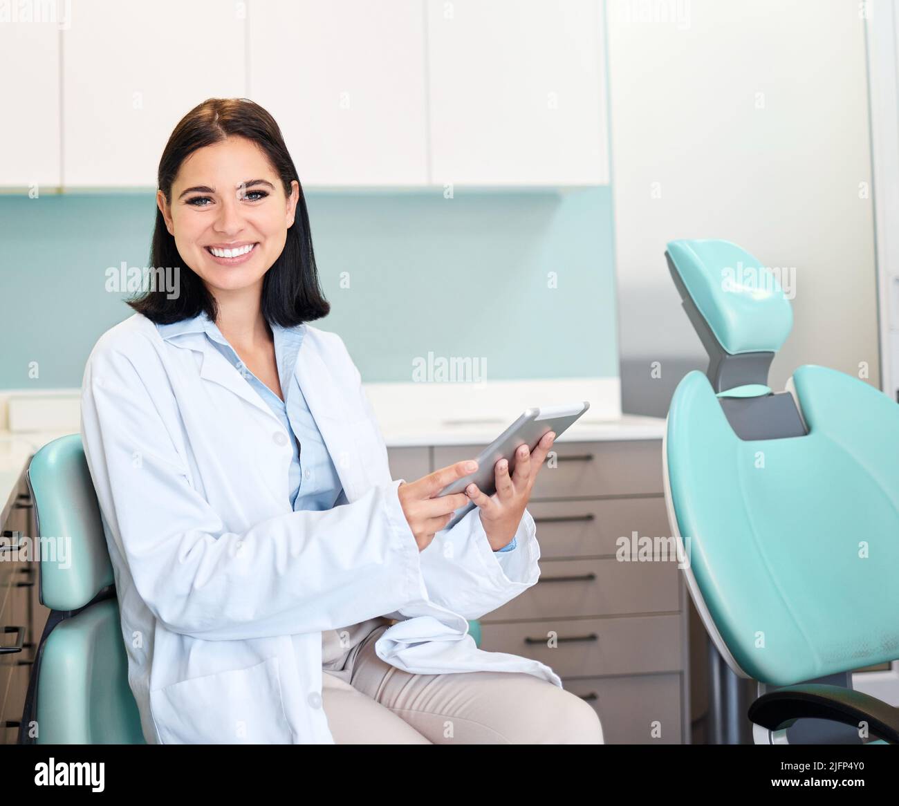 Una bella, sicura dentista femminile seduta nelle sue stanze medici. Una donna sicura e bruna che lavora su un tablet digitale mentre si siede in lei Foto Stock
