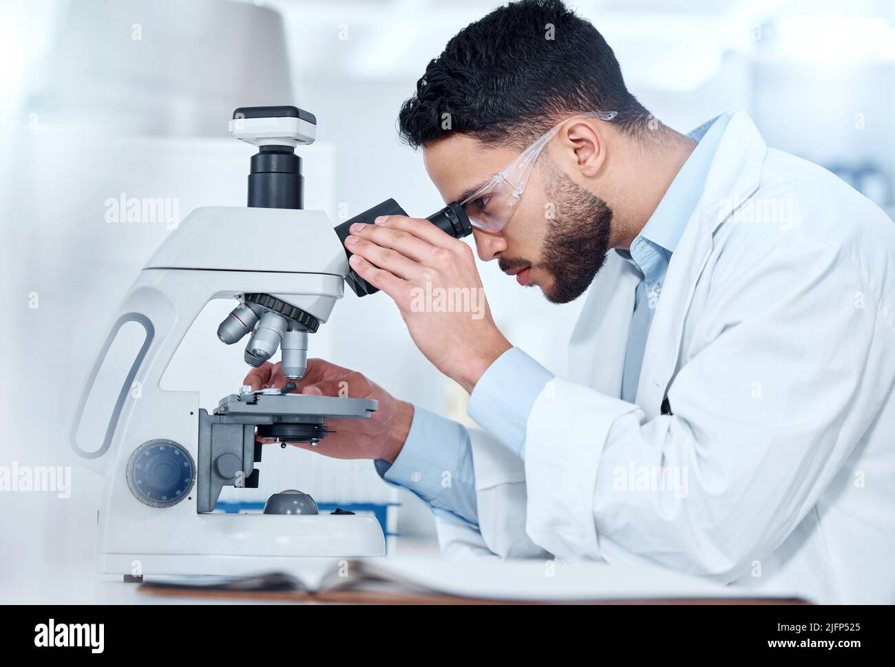 Uno scienziato medico serio seduto su una scrivania e utilizzando un microscopio per esaminare e analizzare campioni di test su vetrini. Professionista sanitario ispanico Foto Stock