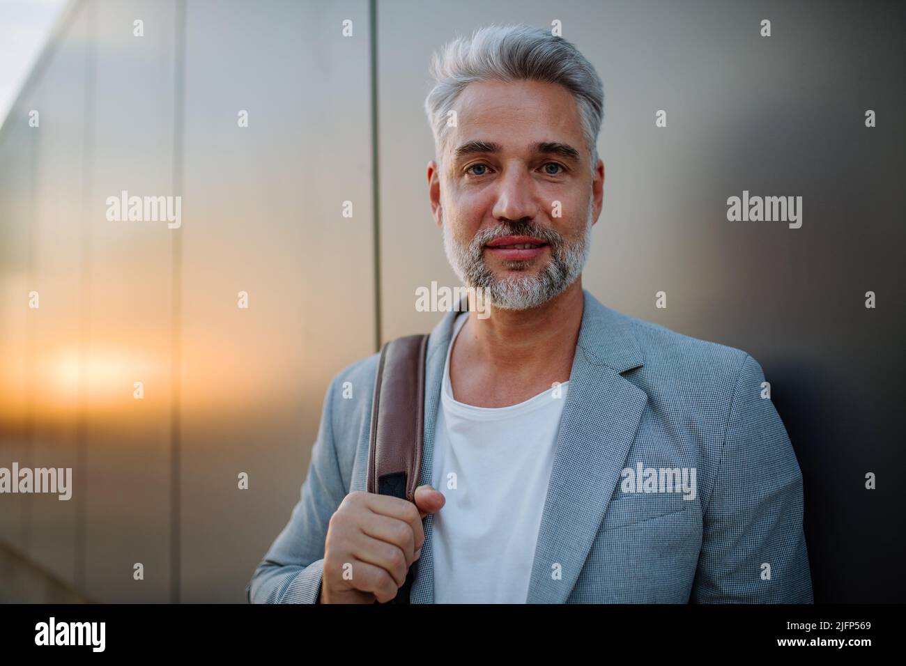 Uomo d'affari maturo e rilassato appoggiandosi sulla parete della strada e guardando la macchina fotografica, concetto di equilibrio della vita lavorativa. Foto Stock