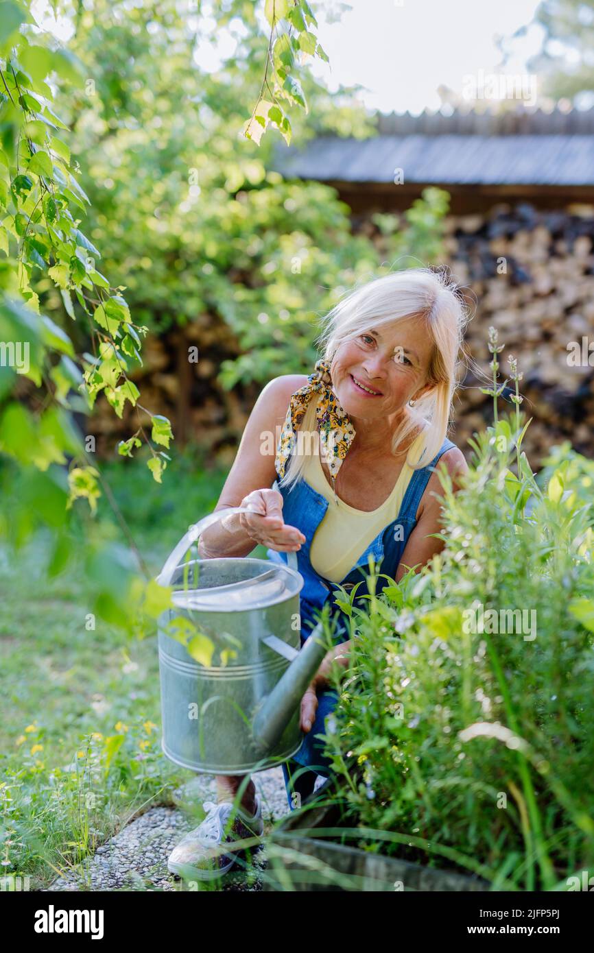 Donna anziana felice che prende la cura dei fiori all'aperto in giardino, innaffiare con la lattina. Foto Stock