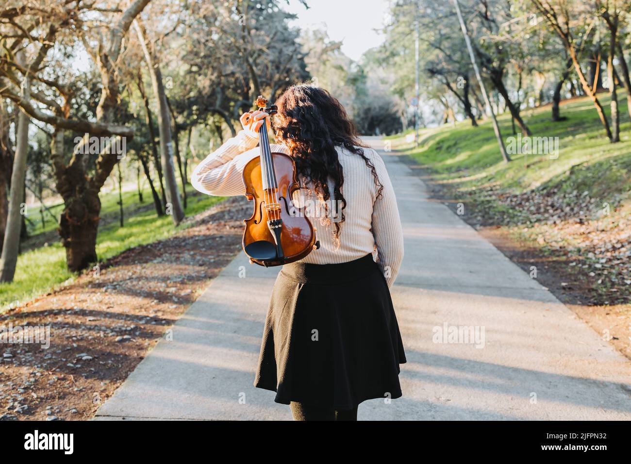 donna con brunette ricurly che tiene un violino sulla schiena, camminando attraverso la strada del parco al tramonto. Foto Stock