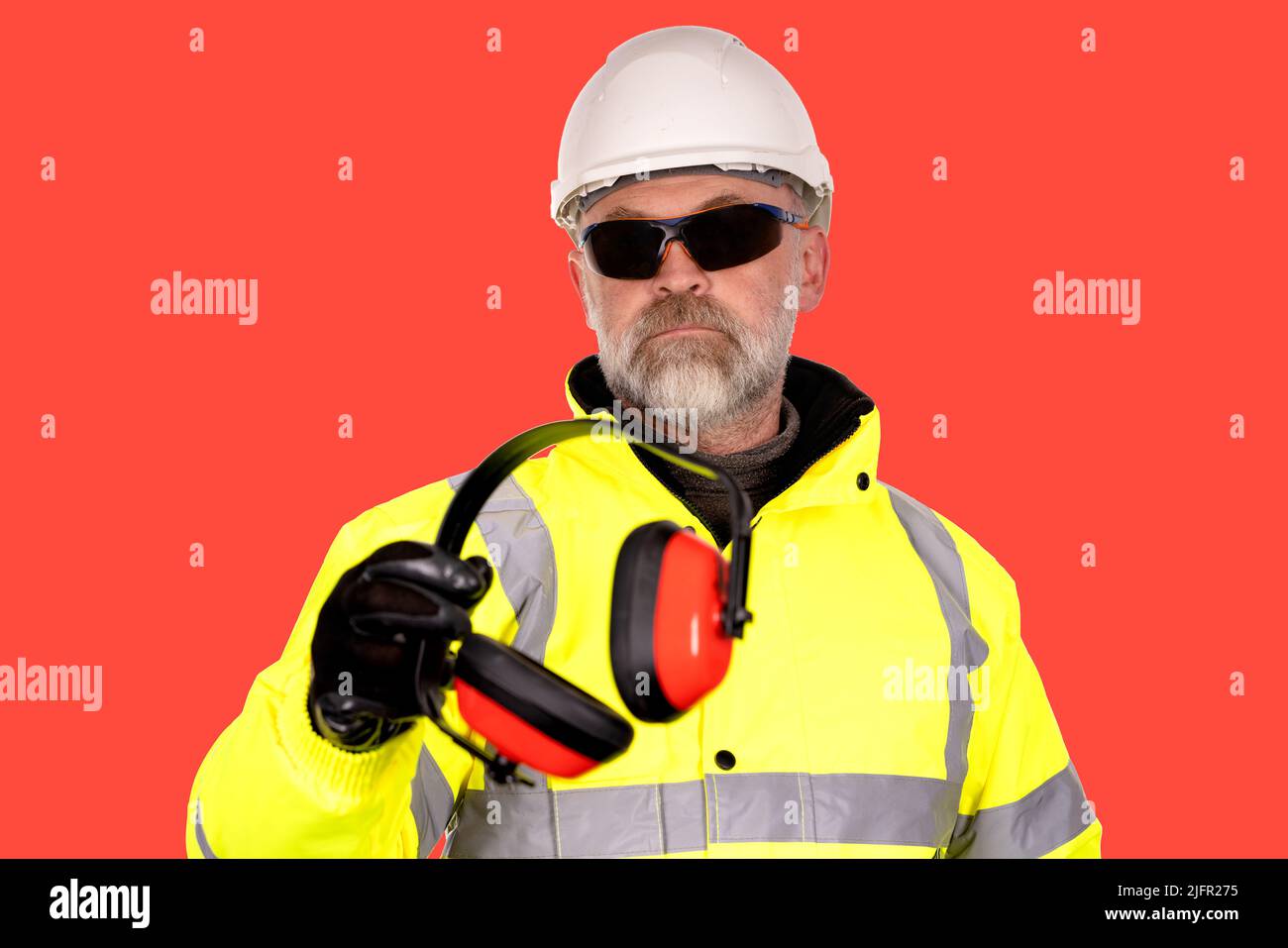 Un operaio di costruzione in un cappello bianco, cappotto giallo hi-viz e occhiali di sicurezza di colore scuro su uno sfondo rosso dona schiuma imbottito cuffie di protezione Foto Stock
