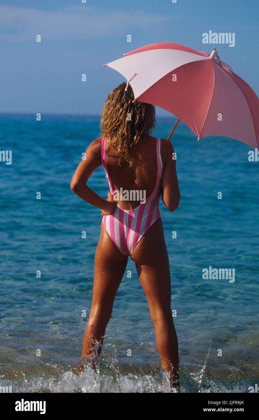 bellezza in piedi sulla spiaggia guardando al mare bella silhouette posteriore con ombrello rosa e costume da bagno blu cielo e acqua di mare backg Foto Stock