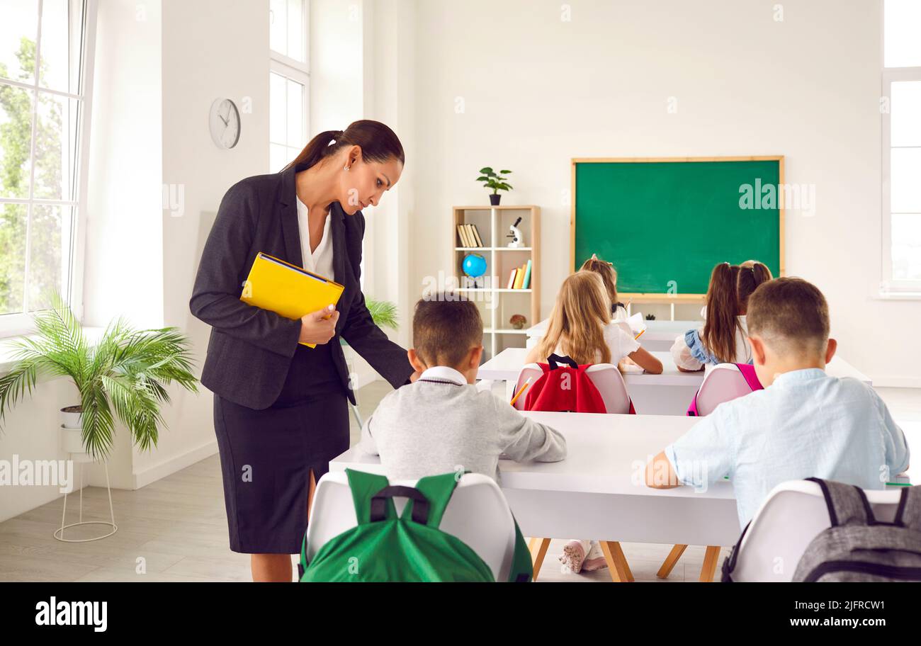 Insegnante di scuola che aiuta i suoi studenti elementari che sono seduti alla scrivania e scrivere Foto Stock