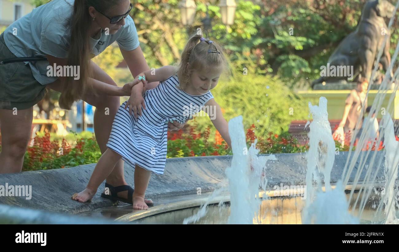 Odessa, Ucraina, Europa dell'Est. 4th luglio 2022. Mamma tiene sorridente bambina giocando con spruzzi d'acqua nella fontana. Mamma con bambina giocare nella fontana nel parco e sorrisi. (Credit Image: © Andrey Nekrasov/ZUMA Press Wire) Foto Stock