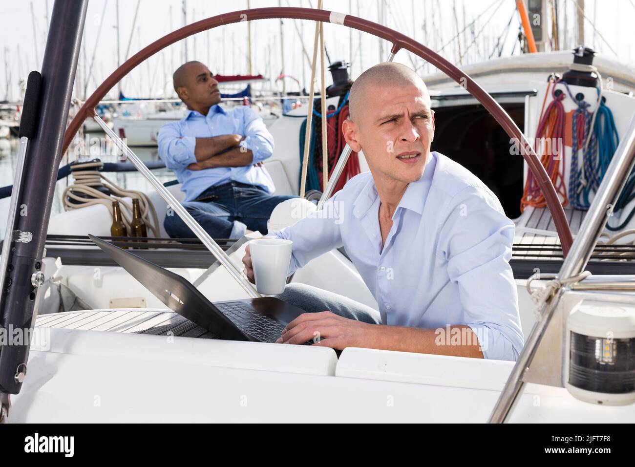 uomo in magliette blu con computer portatile e tazza di caffè su yacht privati in porto al mattino Foto Stock