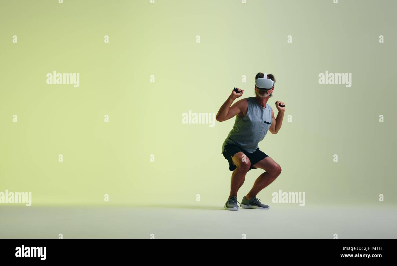 Uomo maturo che solleva un barbell virtuale utilizzando un paio di controller di gioco. Atletico uomo anziano che fa gli esercizi di sollevamento pesi nella realtà virtuale. Senior Foto Stock