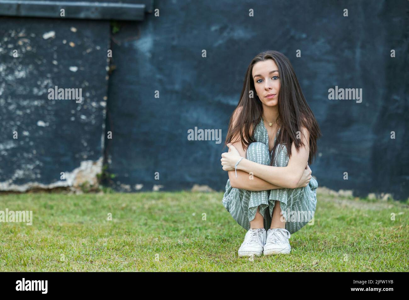 Una bella ragazza bruna teen con uno sguardo serio seduta e appoggiata contro un muro in pensiero con un blu nero ruvido muro della città dietro di lei Foto Stock