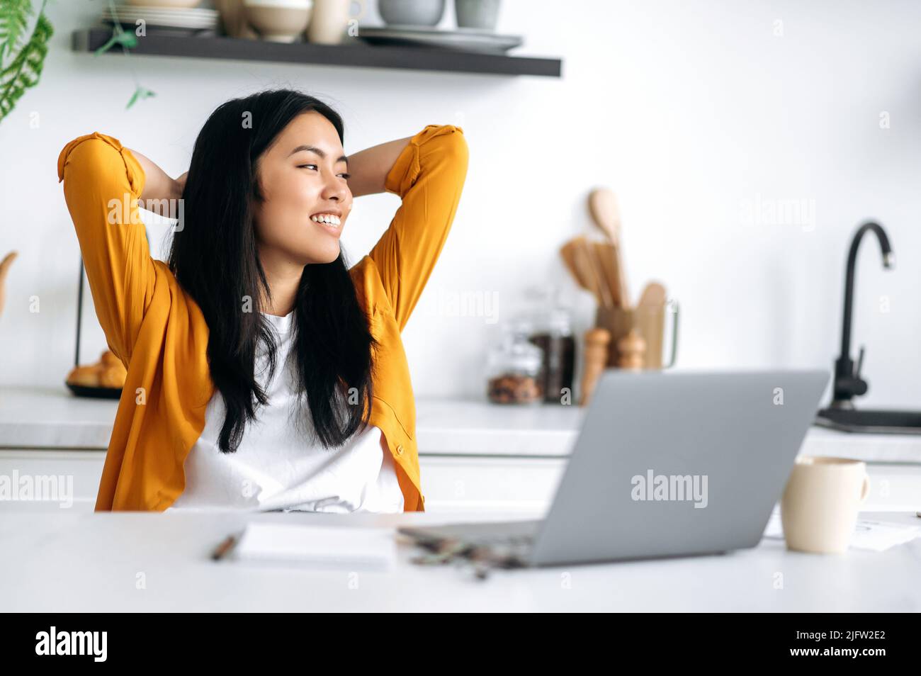 Felice rilassato positivo giovane donna cinese, si siede a un tavolo in cucina, mani incrociate dietro la testa, godendo di tempo di pausa durante il lavoro o lo studio lontano online, guardando da parte, sognando, pianificazione, sorriso Foto Stock