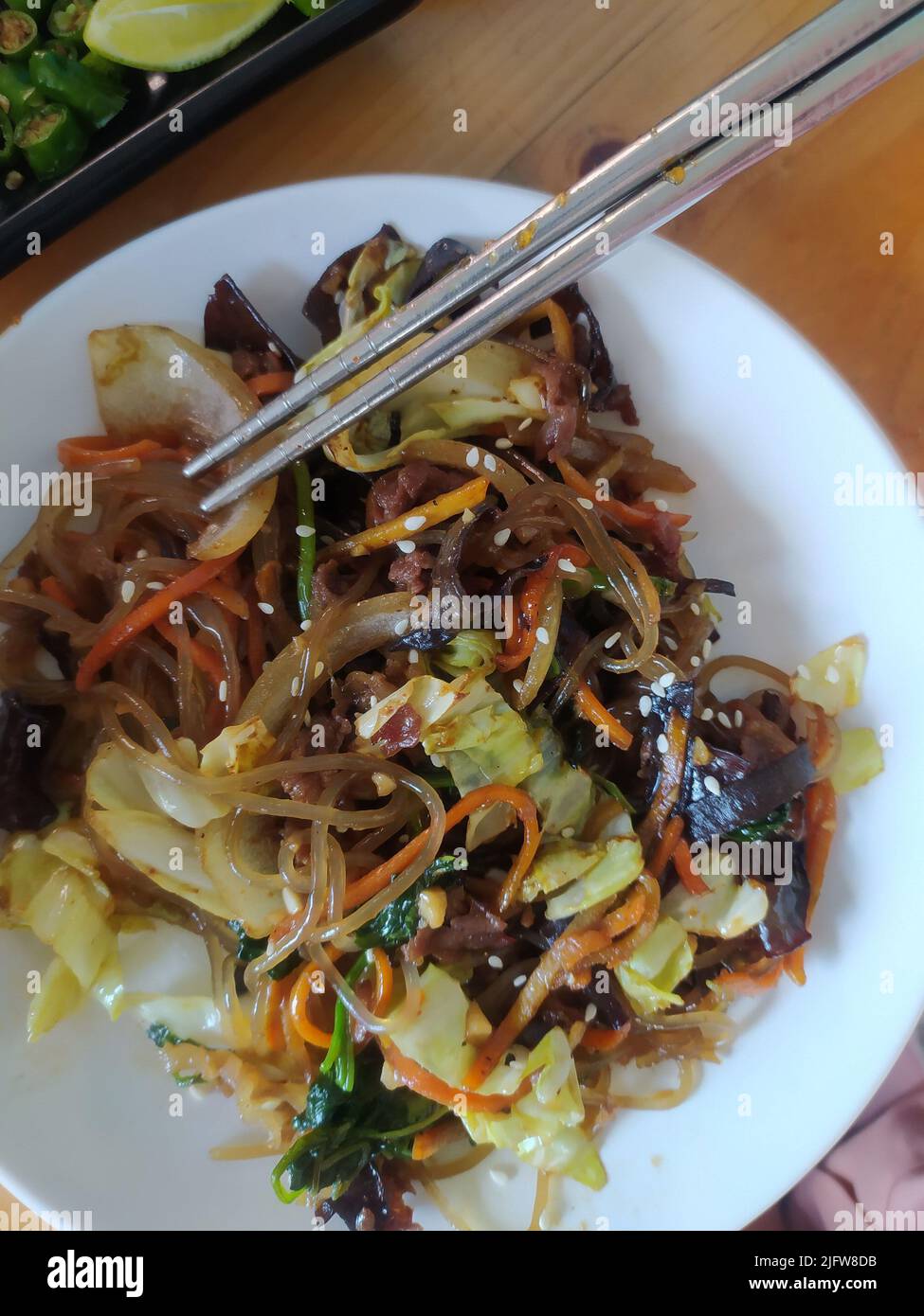 Japchae con carote e semi di sesamo nel piatto bianco sul tavolo. È un piatto salato e leggermente dolce di noodle di vetro e verdure fritte. Foto Stock