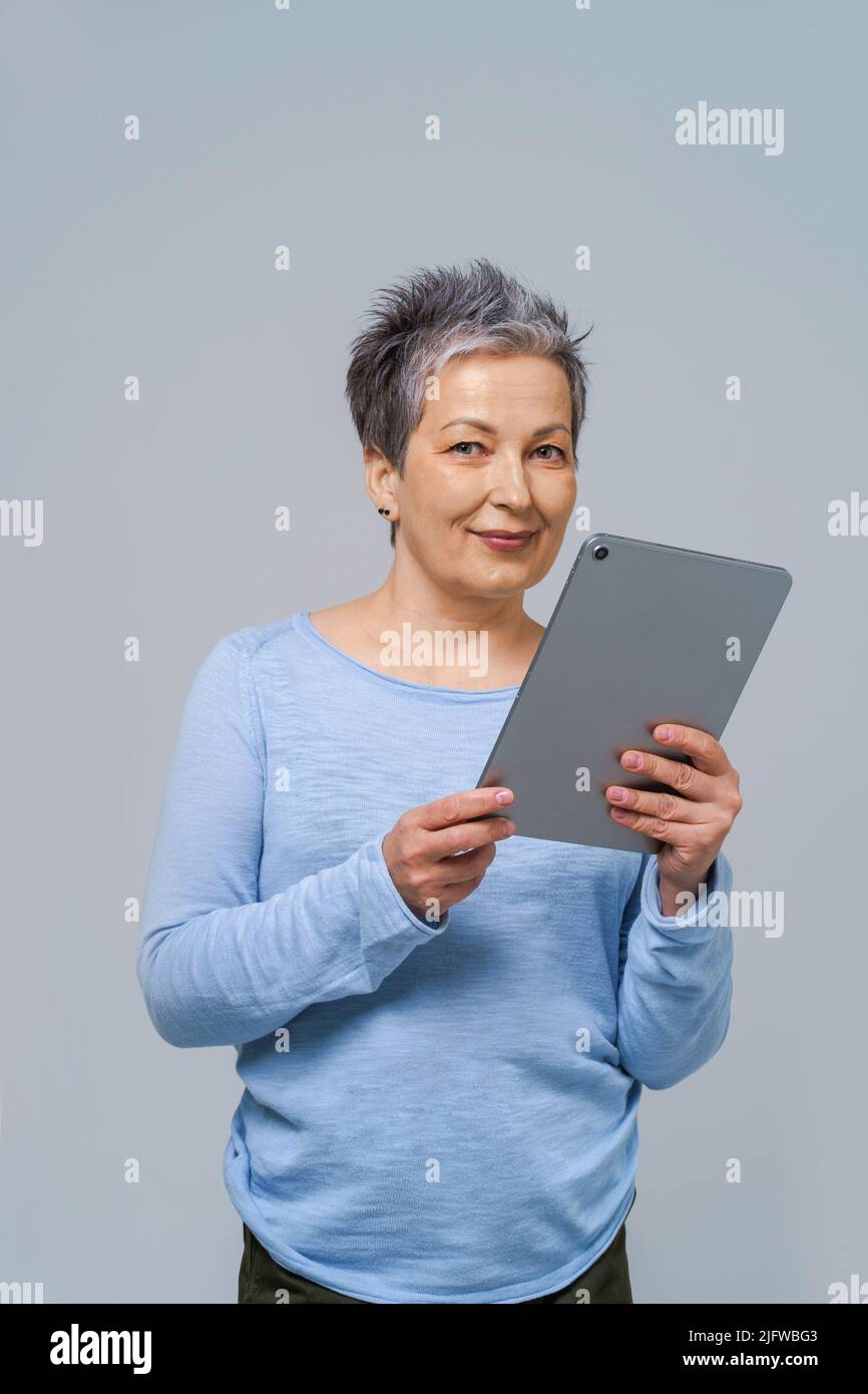 Donna dai capelli grigi con tablet pc in mani che lavorano online. Bella donna nel 50s indossando blusa blu isolato sul grigio. Persone mature e tecnologie. Foto Stock