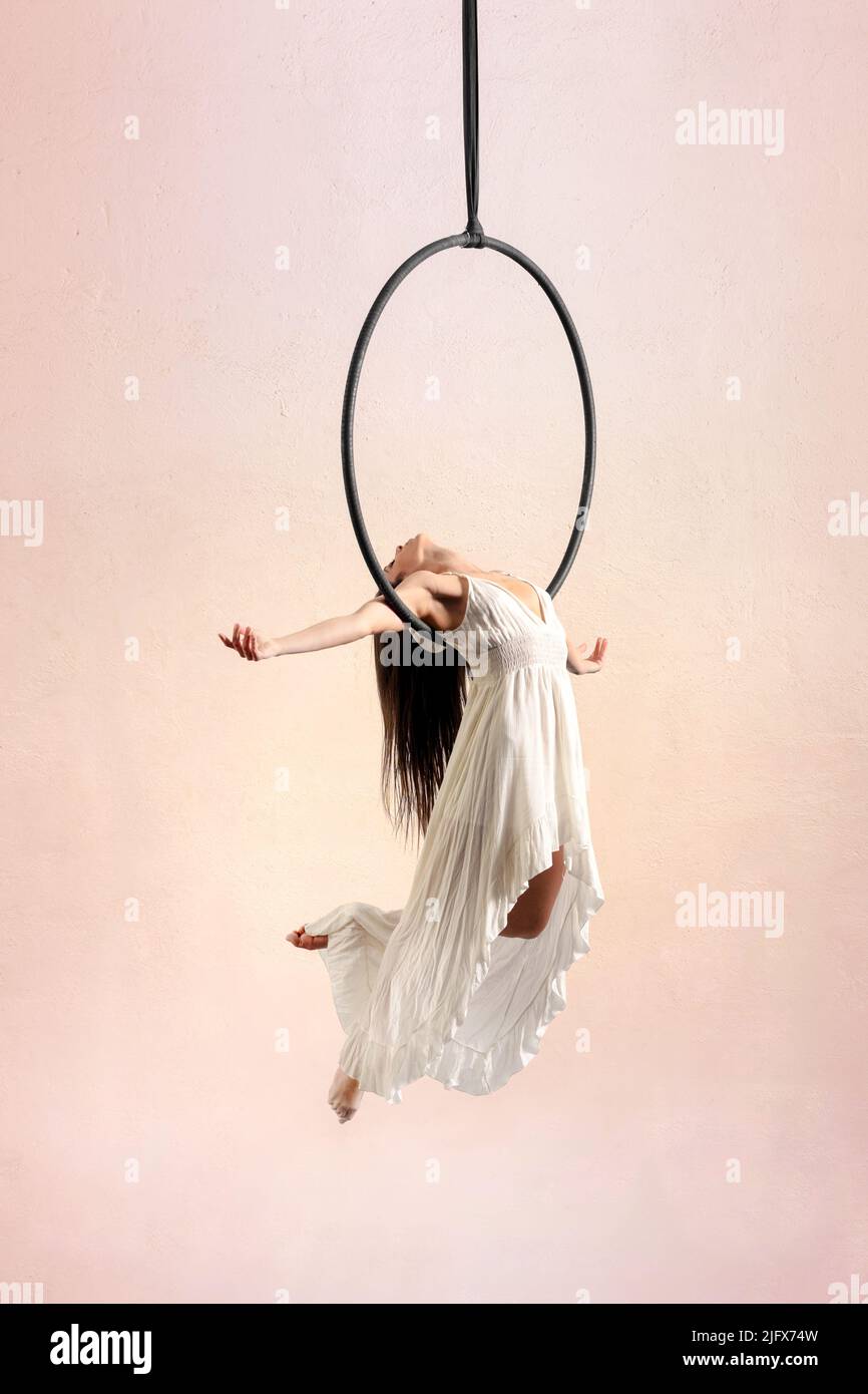 Corpo pieno di aerialista femminile in abito che si esibisce a croce su cerchio aereo durante le prove su sfondo chiaro in studio Foto Stock