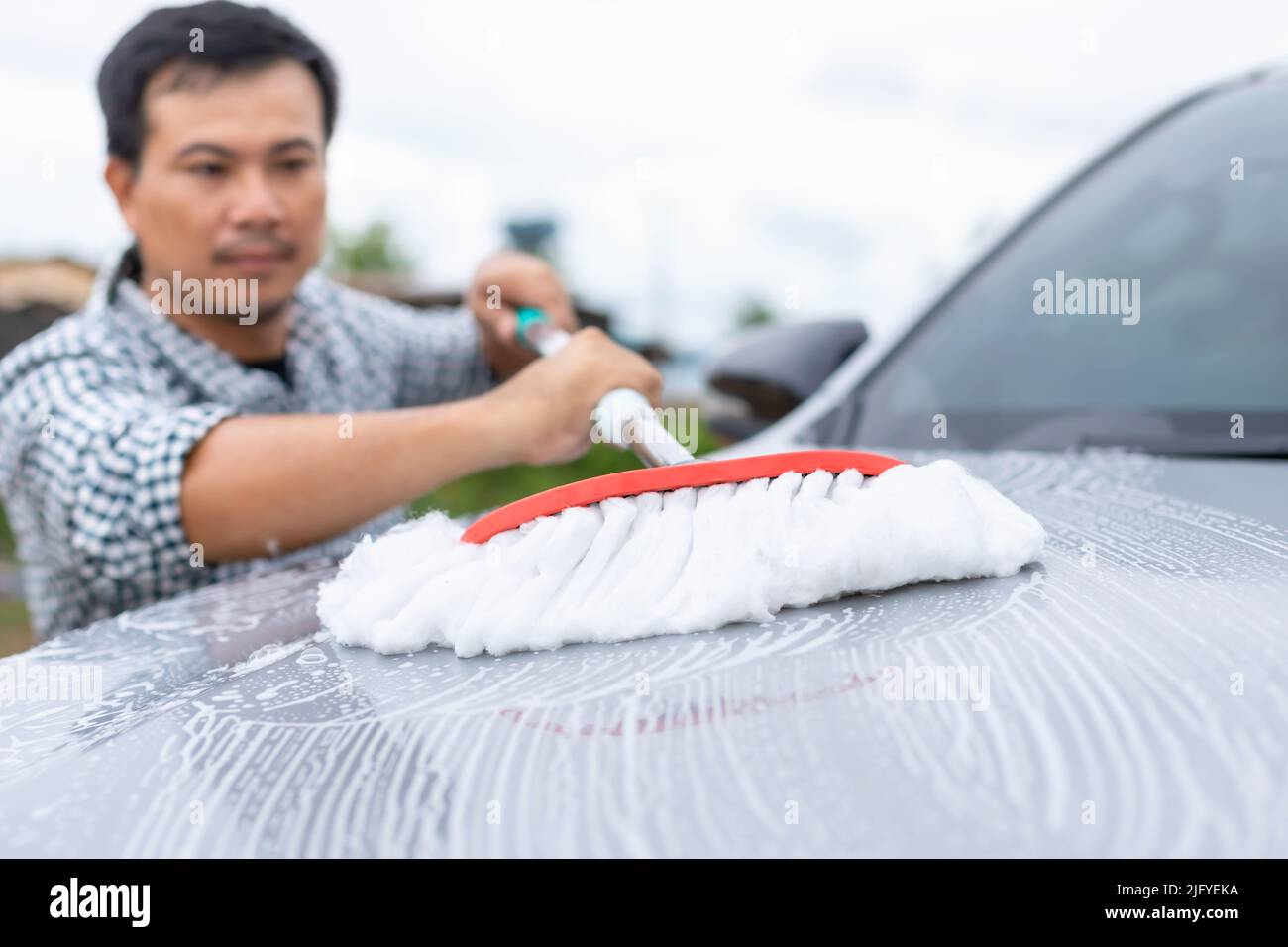 Uomo asiatico utilizzando mop bianco con sapone per lavare l'auto all'aperto. Concetto di pulizia e manutenzione dell'auto Foto Stock