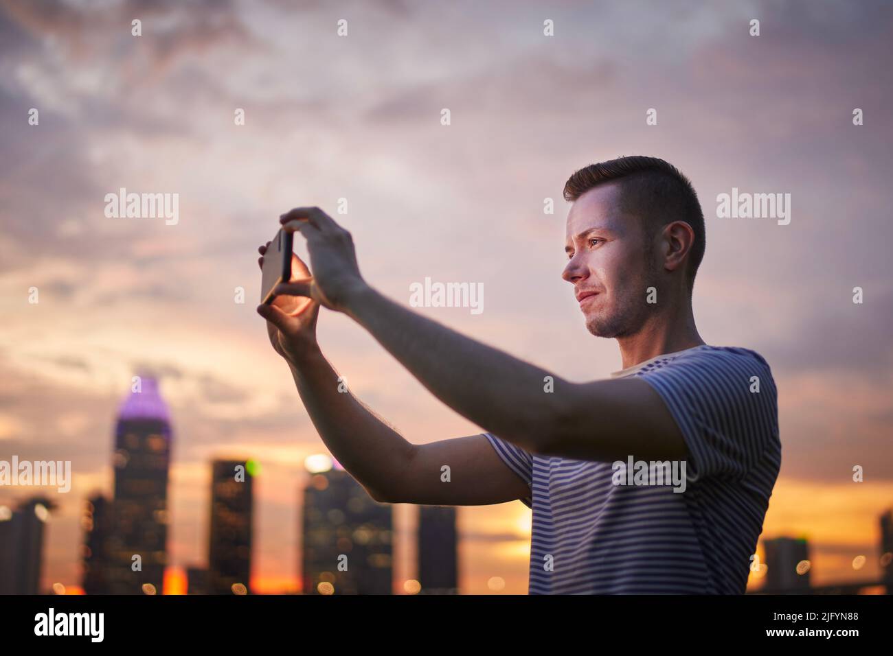 Uomo che scatta foto tramite telefono cellulare dello skyline urbano al tramonto d'oro, Singapore. Foto Stock