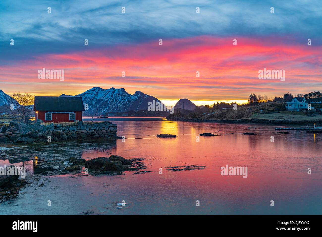 Cielo di Fiery all'alba su un Rorbu rosso di fronte al mare congelato, Ballstad, Vestvagoy, Isole Lofoten, Norvegia, Scandinavia, Europa Foto Stock