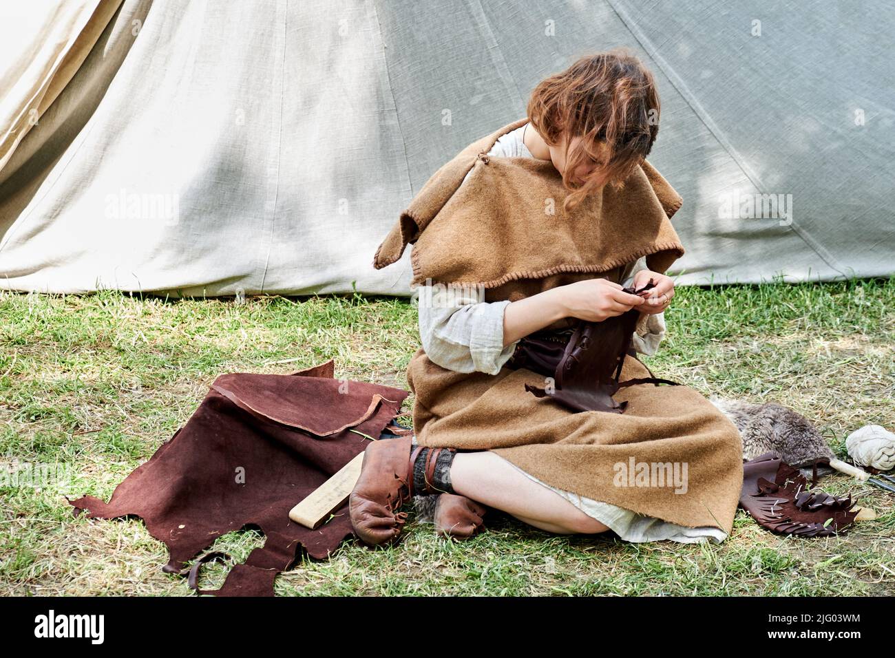 Una donna in vecchi abiti romani sews pelle seduta sul prato. Ricostruzione degli eventi dell'Impero Romano. Festival di tempi ed epoche sulla str Foto Stock