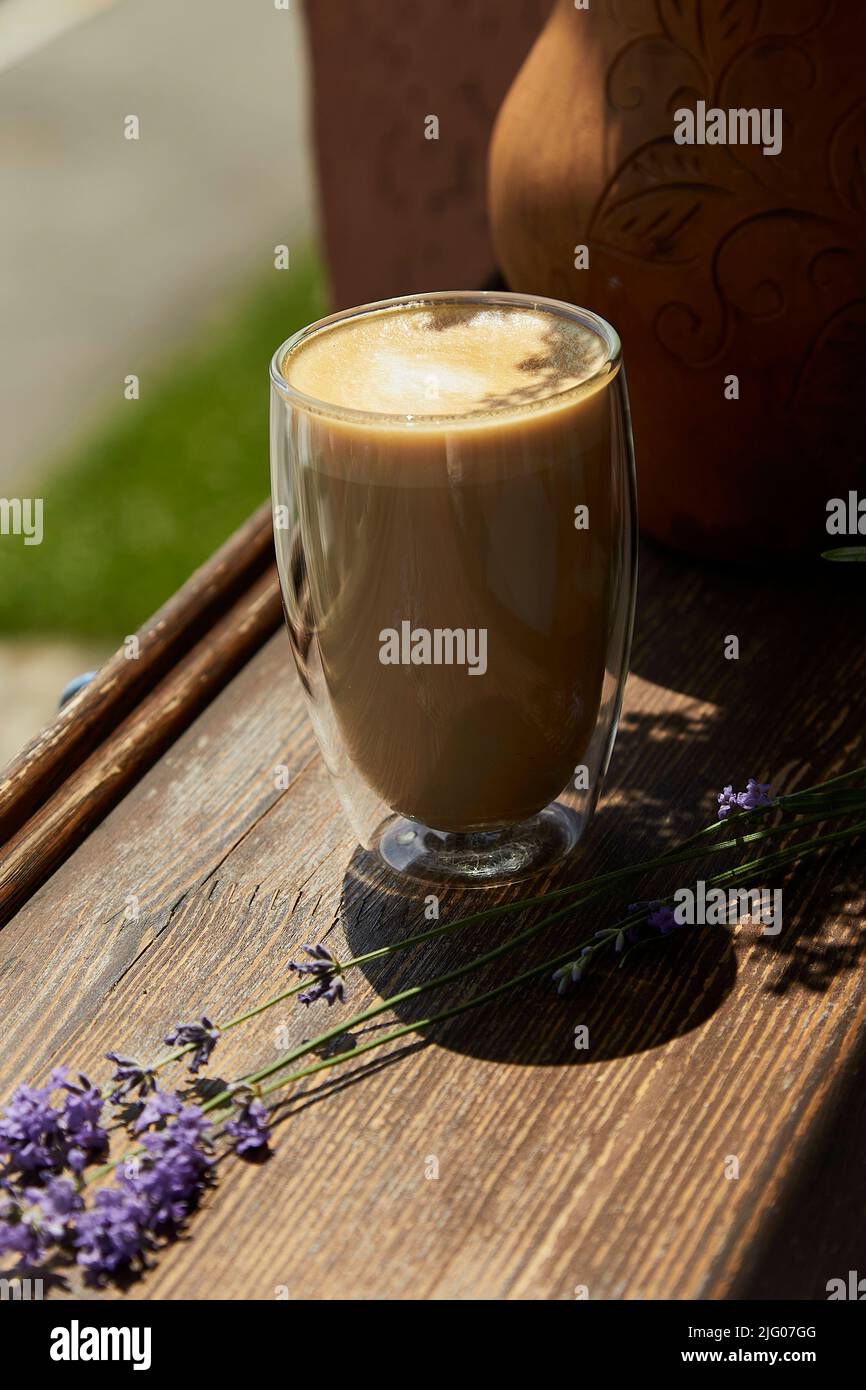 Vetro con doppio fondo di latte su tavola di legno sotto ombre dure. Decorazioni di fiori di lavanda all'esterno nella terrazza del caffè. Pausa caffè atmosferica Foto Stock