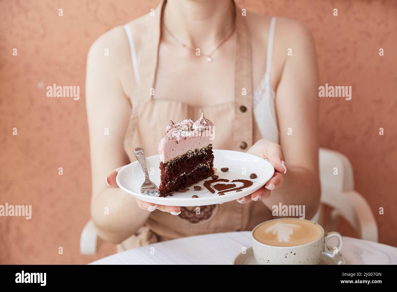 La donna tiene un pezzo di torta al cioccolato alla frutta e una tazza di cappuccino su un tavolo di legno all'esterno nella terrazza del caffè. Caffè d'atmosfera, tempo per te. Decorazioni a forma di cuore per dessert Foto Stock