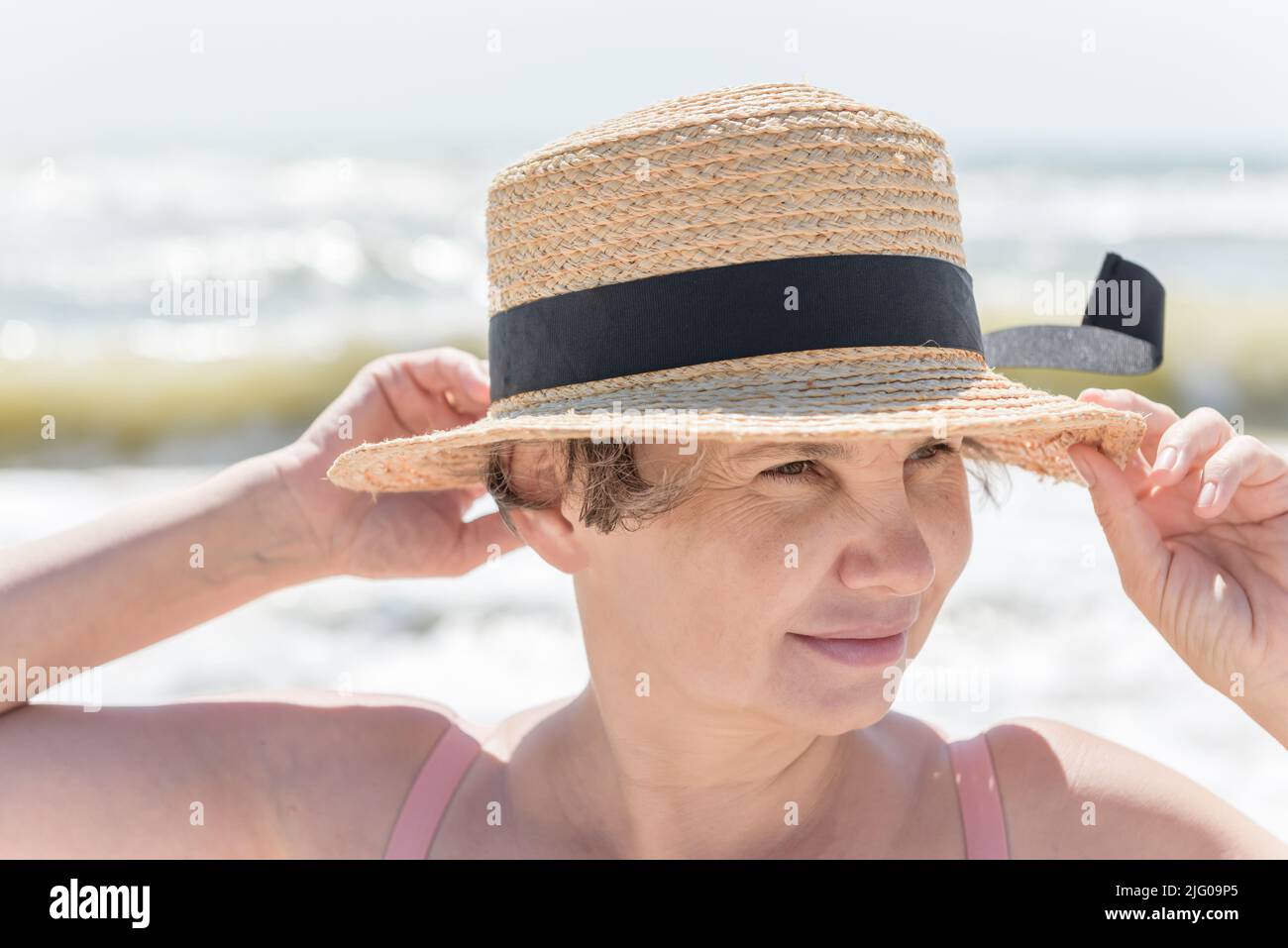 Donna dai capelli grigi con un lieve sorriso che sostiene i bordi del suo cappello, che vento dal mare cercando di strappare la testa, guardando qualcosa sulla riva. Foto Stock