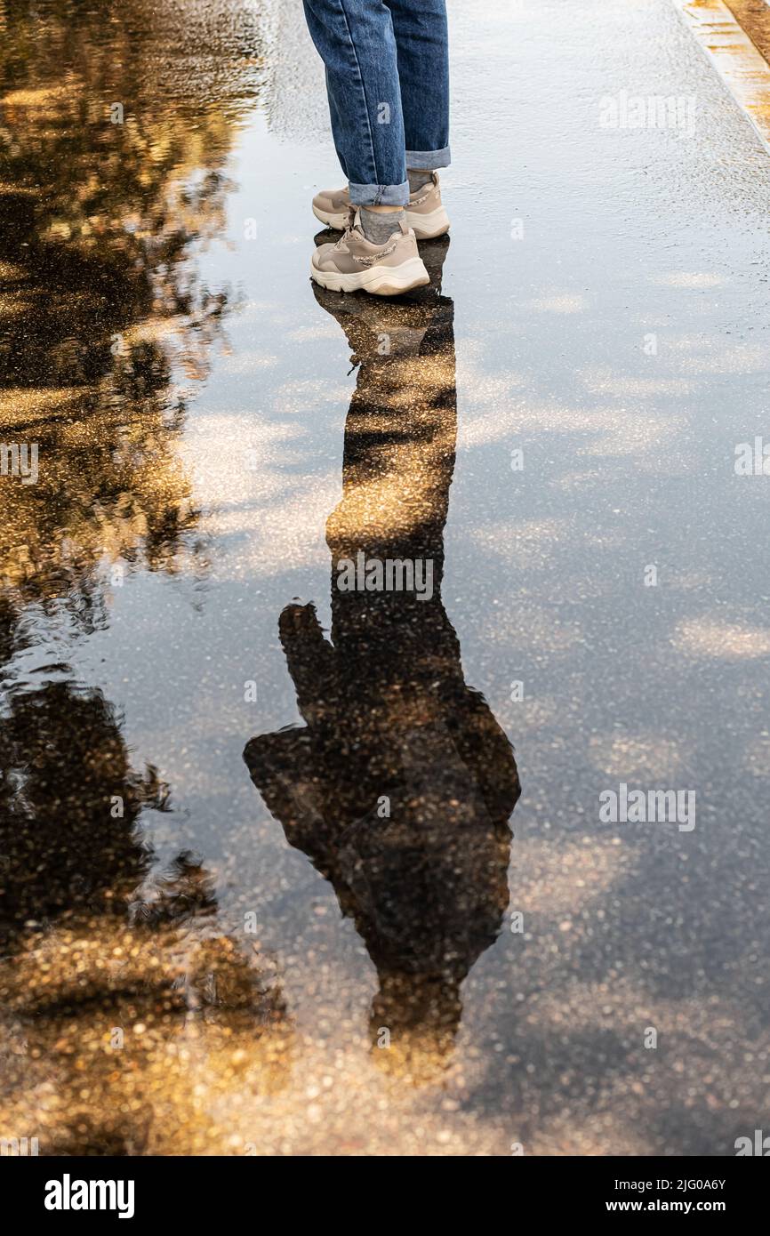 Gambe di donna in jeans e sneakers. La silhouette di una donna si riflette in una pozzanghera sul marciapiede. Cammina dopo la pioggia. Foto Stock