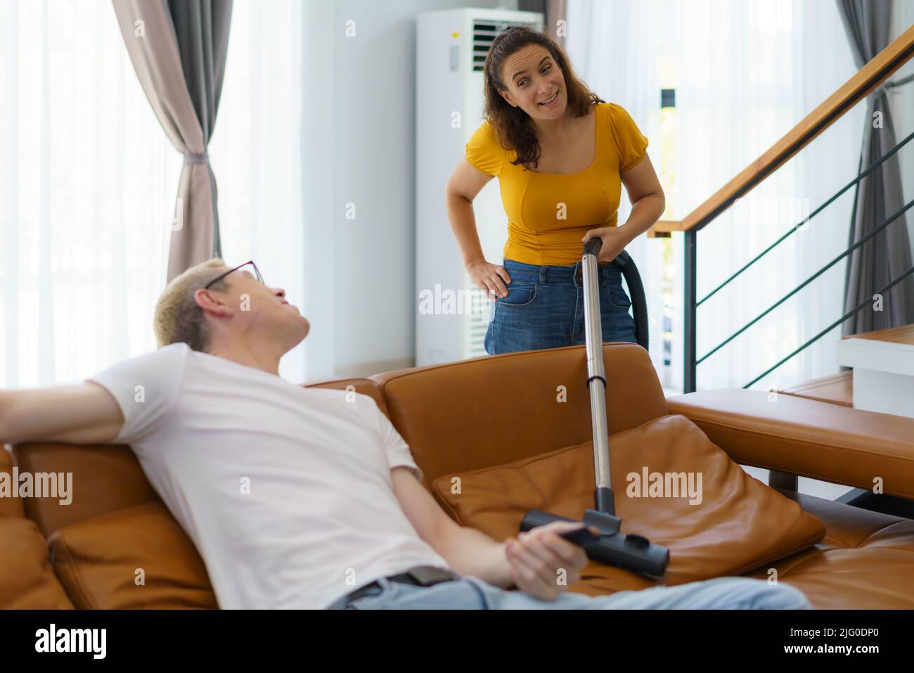 La moglie caucasica che usa l'aspirapolvere nel soggiorno a casa sconvolta suo marito per non aiutare con il lavoro domestico e sedersi comodamente e guardare la TV. Foto Stock