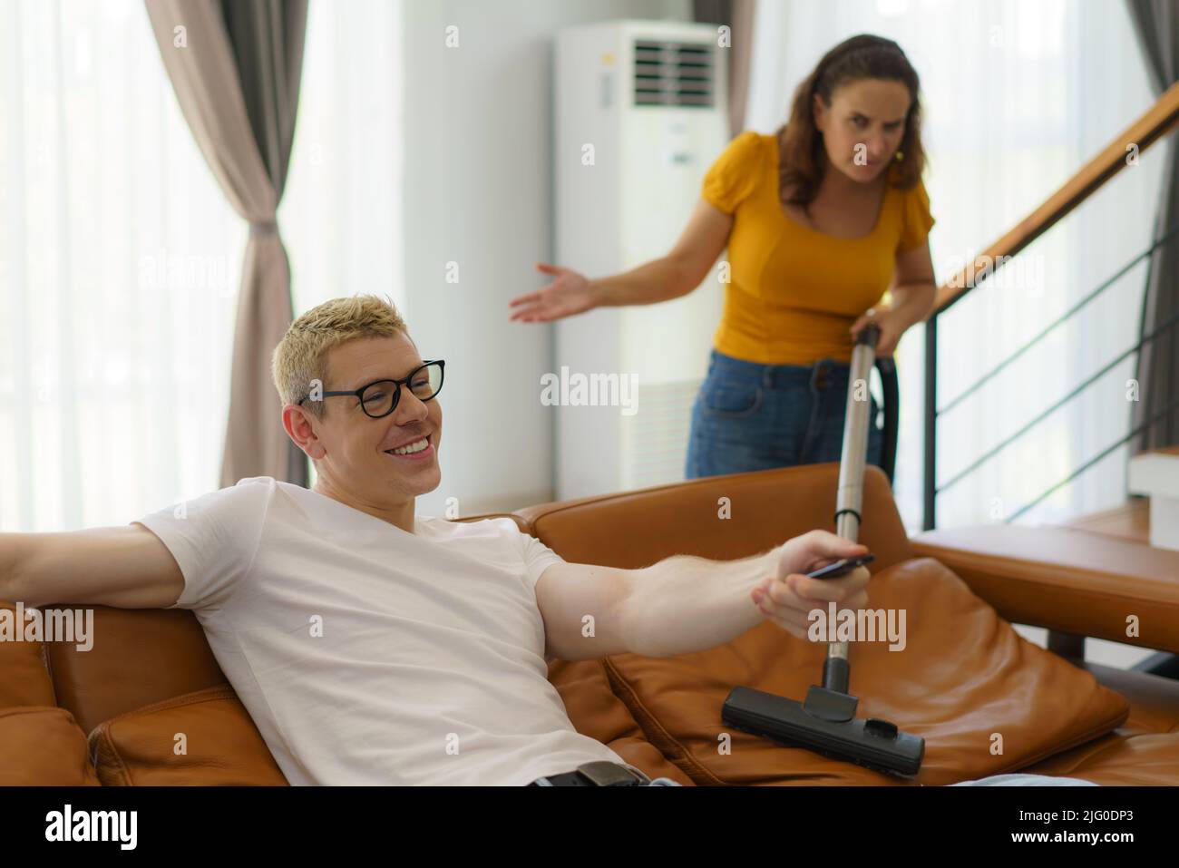 La moglie caucasica che usa l'aspirapolvere nel soggiorno a casa sconvolta suo marito per non aiutare con il lavoro domestico e sedersi comodamente e guardare la TV. Foto Stock