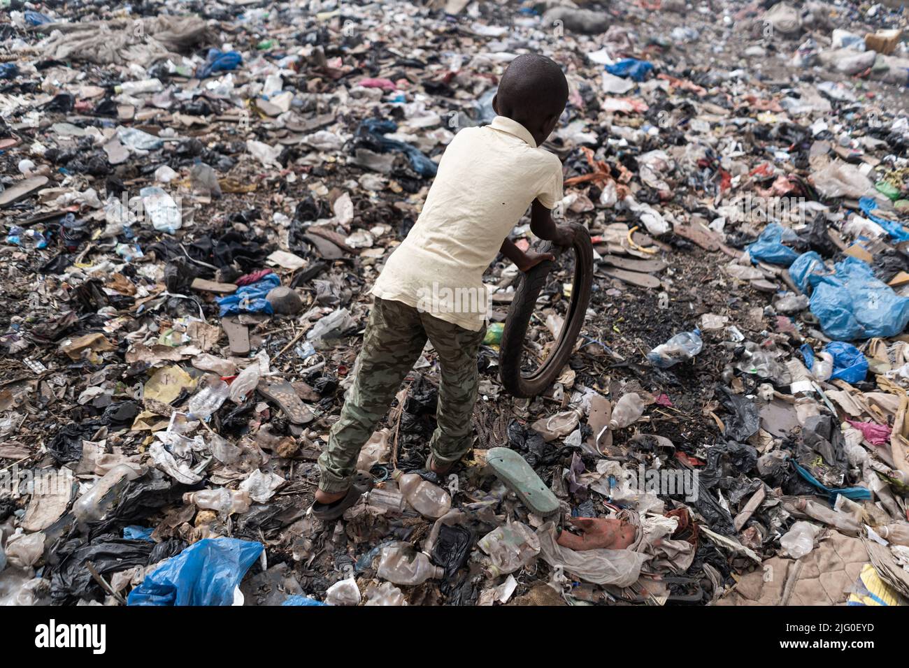 Vista dall'alto di un piccolo ragazzo africano che gioca con un vecchio pneumatico da bicicletta in una zona fortemente inquinata; simbolo dell'infanzia nei paesi sottosviluppati Foto Stock