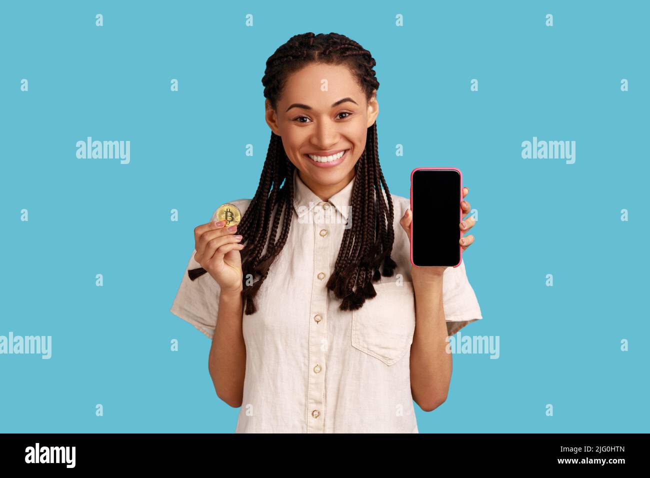 Bella donna con dreadlocks nero sorridente e tenendo il telefono cellulare e bitcoin d'oro, pubblicità di applicazioni cripto, indossare camicia bianca. Studio interno girato isolato su sfondo blu. Foto Stock