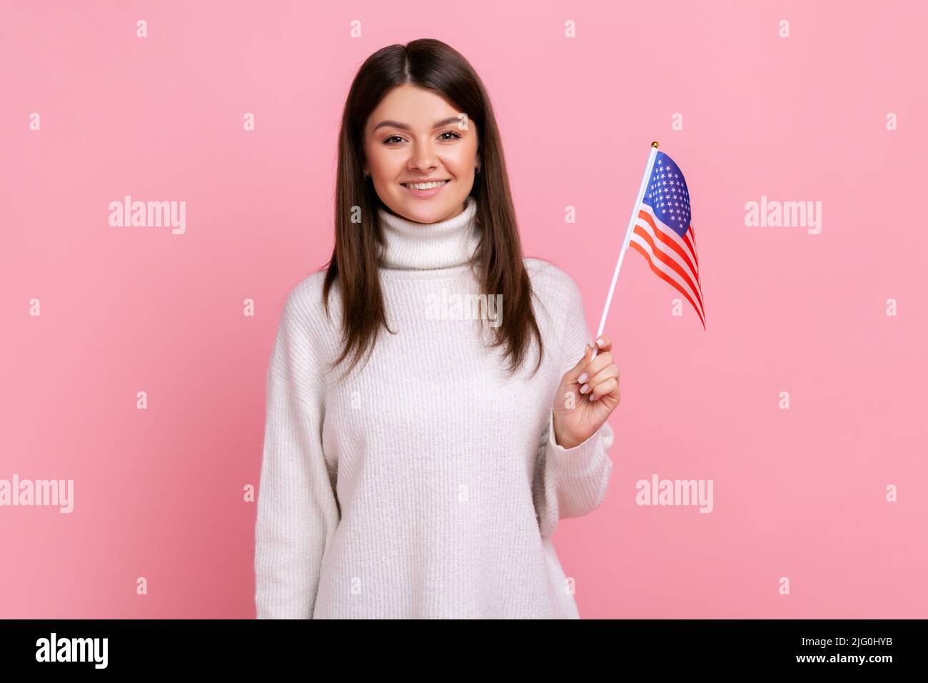 Donna bruna attraente che tiene la bandiera americana in mano e guarda la macchina fotografica con positivo, indossando bianco casual stile maglione. Studio interno girato isolato su sfondo rosa. Foto Stock