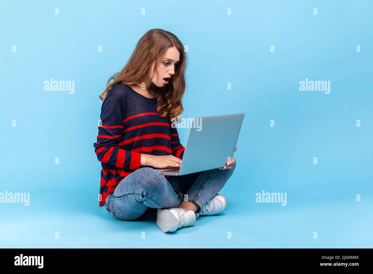 Donna stupita che indossa un pullover casual a righe, seduta sul pavimento con gambe incrociate e guardando lo schermo del laptop con espressione scioccata. Studio interno girato isolato su sfondo blu. Foto Stock