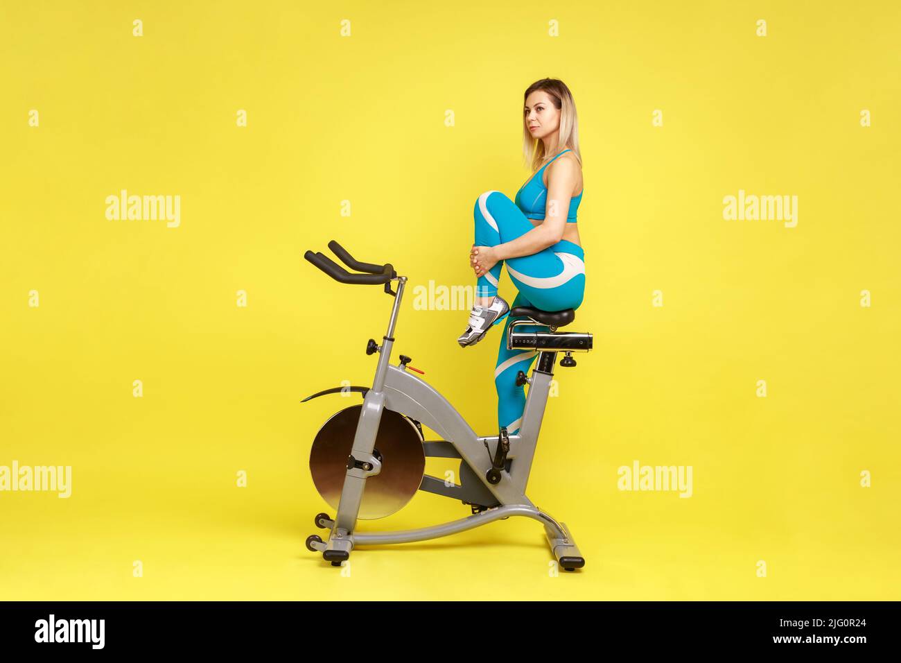 Ritratto della vista laterale di attraente istruttore donna che allunga la gamba prima di allenarsi in bicicletta, guardando via, indossando abbigliamento sportivo blu. Studio interno girato isolato su sfondo giallo. Foto Stock