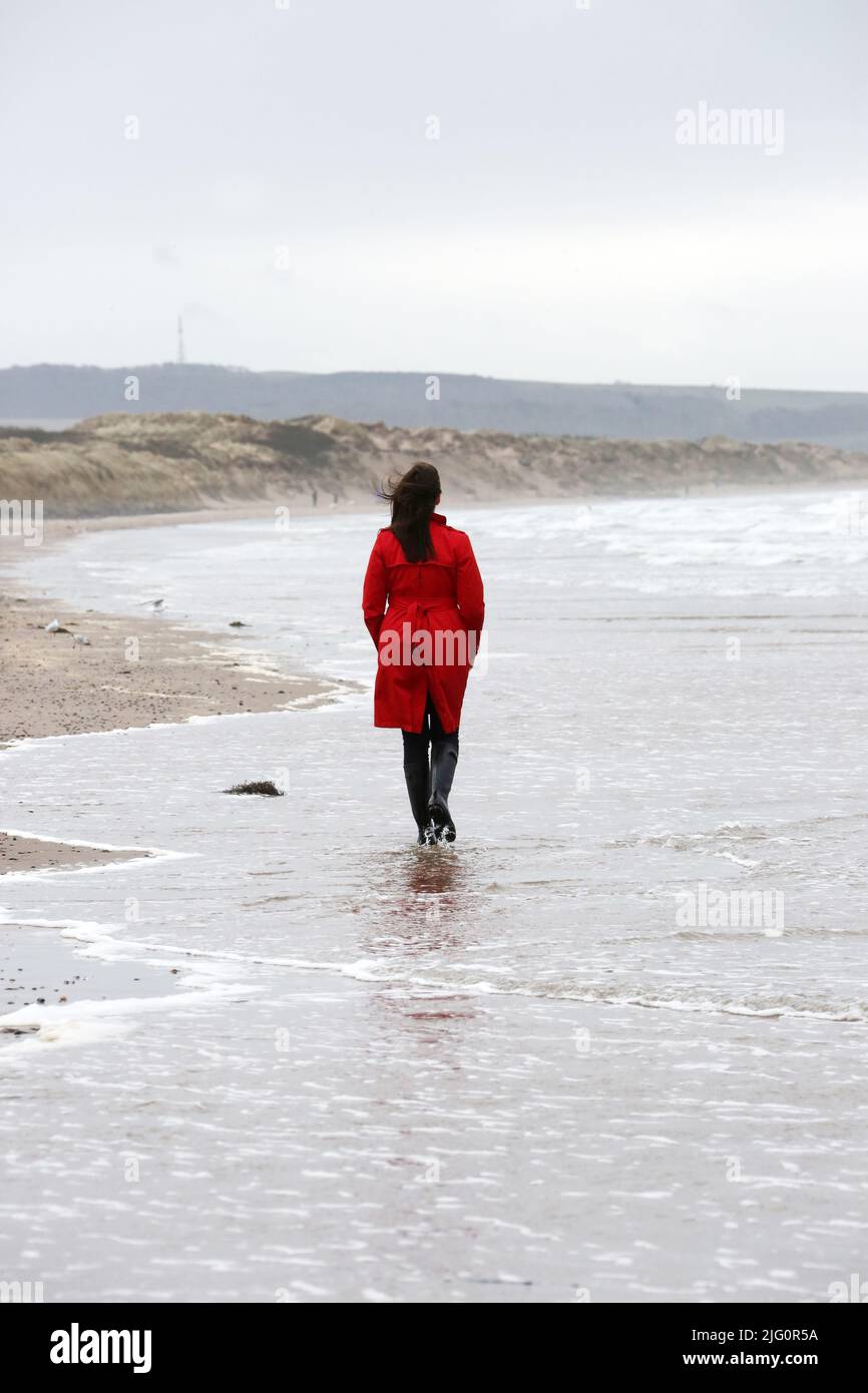 Donne cauasiche bianche di età media che indossano un cappotto rosso trenchcoat , wellingtons neri da soli su spiaggia bagnata e ventosa come le onde si rompono, isolate, premurose, enigmatiche con spazio per la copia e il testo Foto Stock