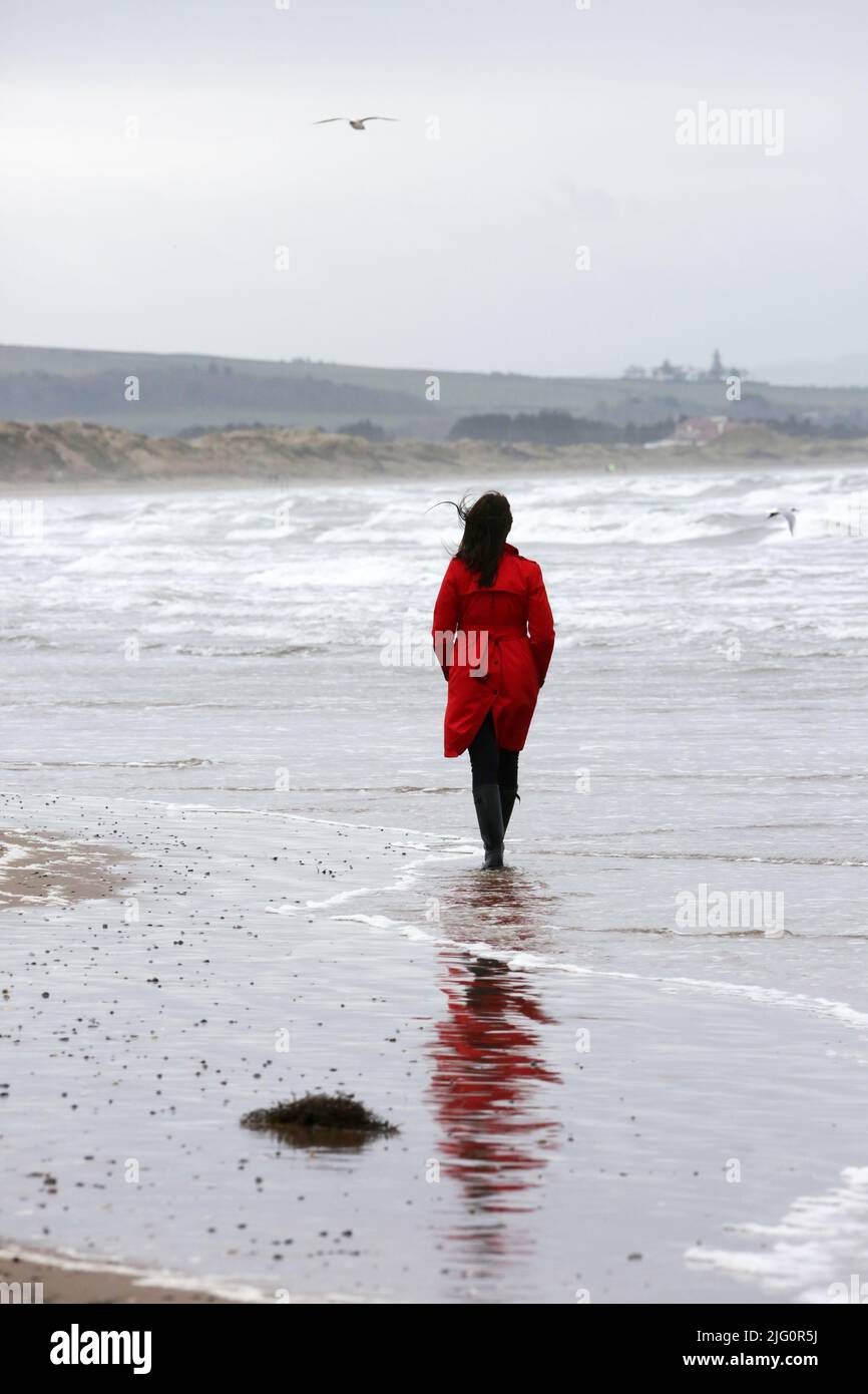 Donne cauasiche bianche di età media che indossano un cappotto rosso trenchcoat , wellingtons neri da soli su spiaggia bagnata e ventosa come le onde si rompono, isolate, premurose, enigmatiche con spazio per la copia e il testo Foto Stock