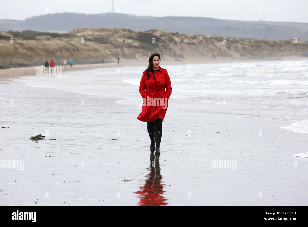 Donne cauasiche bianche di età media che indossano un cappotto rosso trenchcoat , wellingtons neri da soli su spiaggia bagnata e ventosa come le onde si rompono, isolate, premurose, enigmatiche con spazio per la copia e il testo Foto Stock