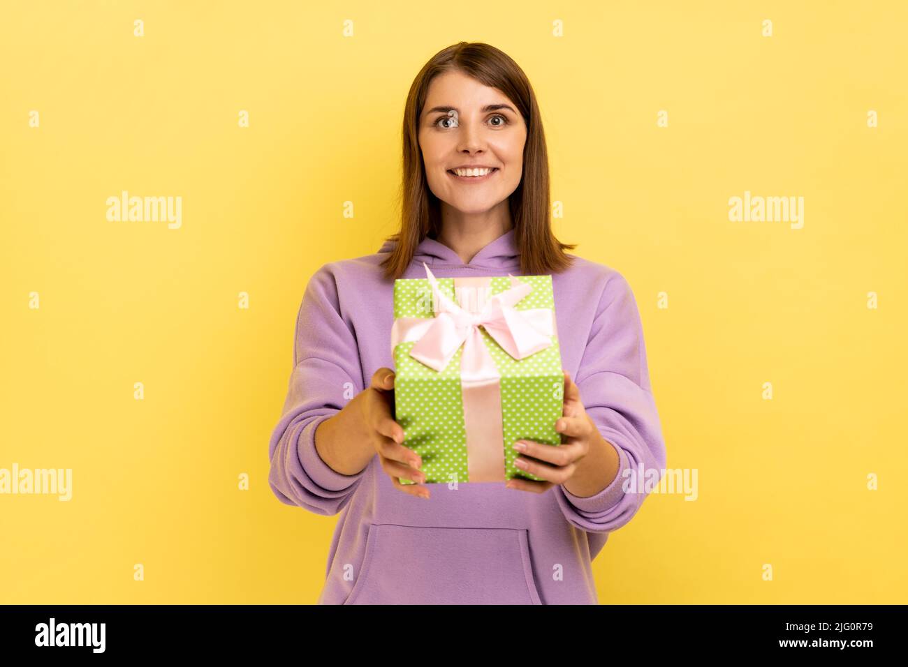 Divertente Happy donna dai capelli scuri dando scatola blu alla macchina fotografica e sorridendo, condividendo regalo di festa, congratulandosi per il compleanno, indossando felpa con cappuccio viola. Studio interno girato isolato su sfondo giallo. Foto Stock