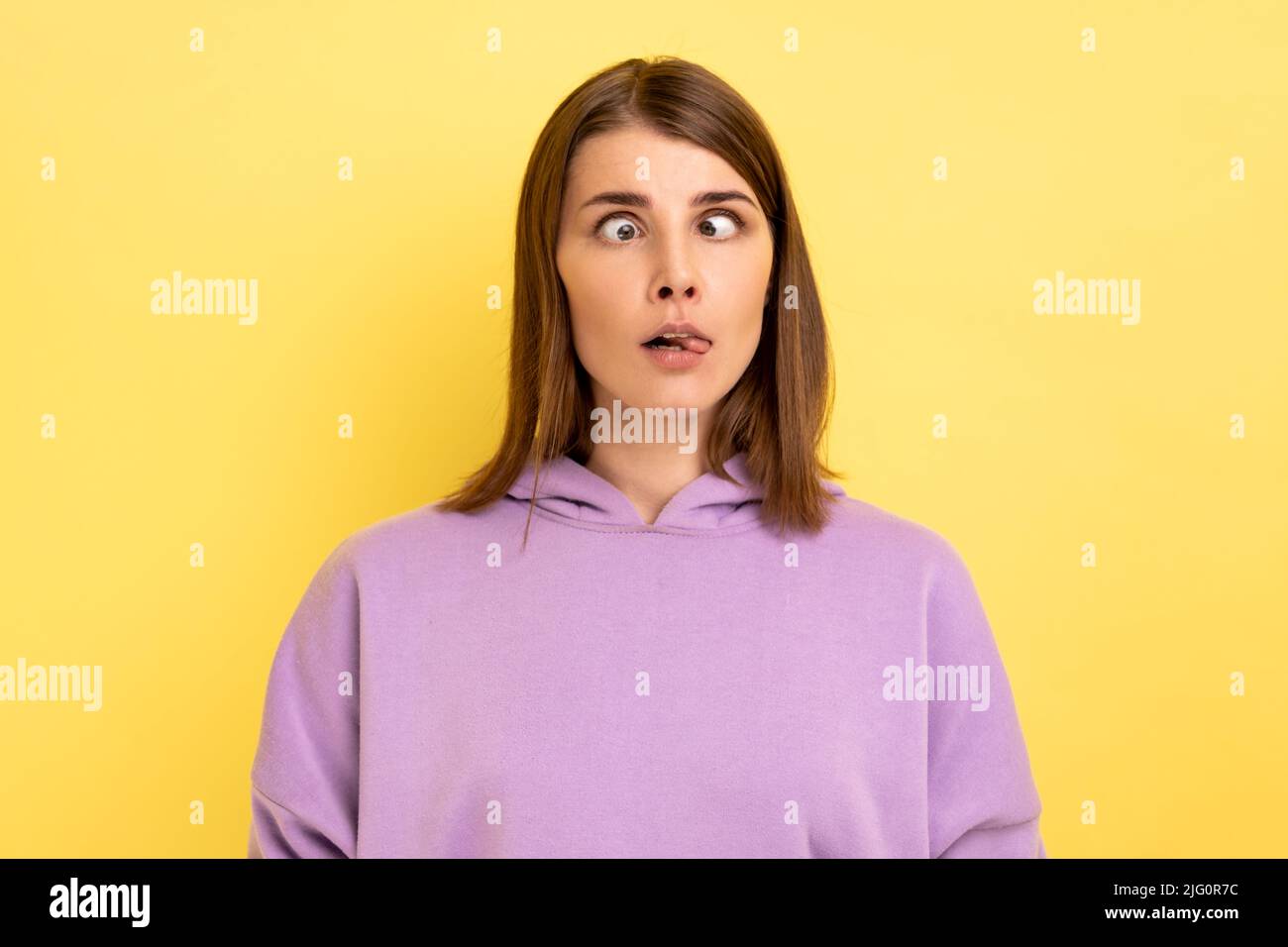 Ritratto di giovane donna adulta stolita con capelli in piedi con occhi incrociati e lingua fuori, comportamento infantile, indossando felpa con cappuccio viola. Studio interno girato isolato su sfondo giallo. Foto Stock