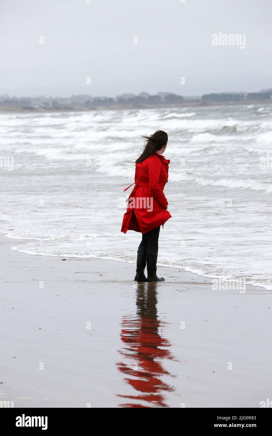 Donne cauasiche bianche di età media che indossano un cappotto rosso trenchcoat , wellingtons neri da soli su spiaggia bagnata e ventosa come le onde si rompono, isolate, premurose, enigmatiche con spazio per la copia e il testo Foto Stock